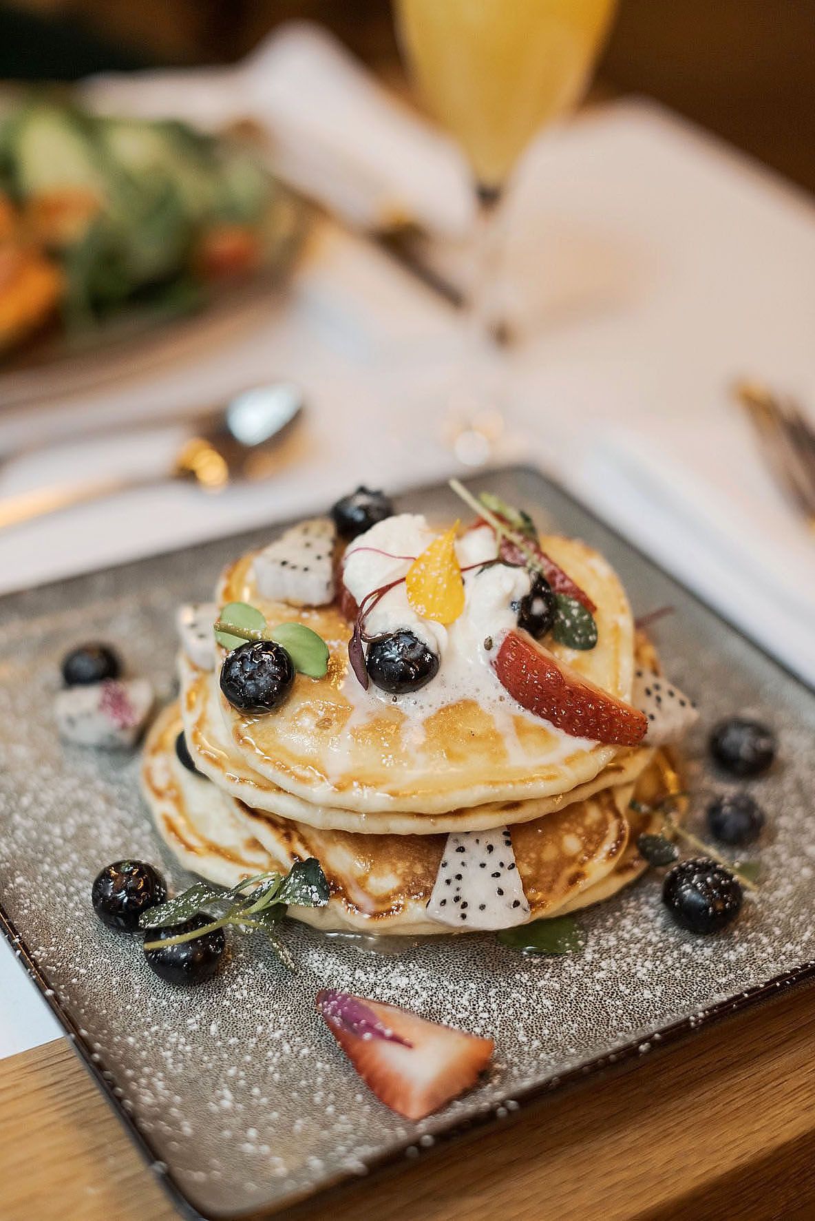 Stack of fluffy pancakes topped with fresh blueberries, dragon fruit, peaches and whipped cream served on gray plate.