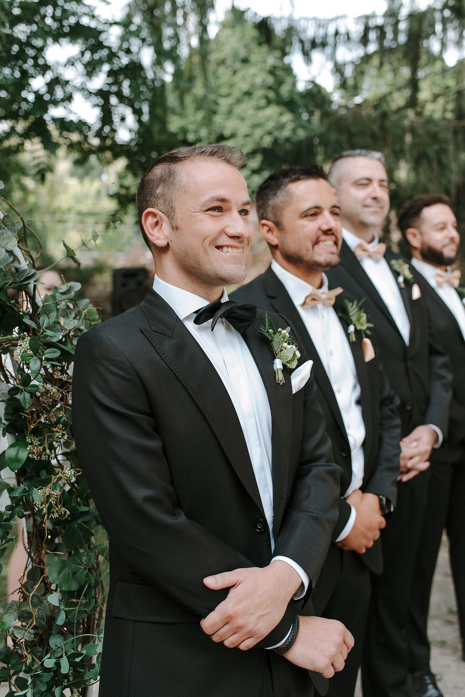 Groomsmen in black tuxedos and bowties stand together outdoors with greenery in the background on wedding day. La Toundra.