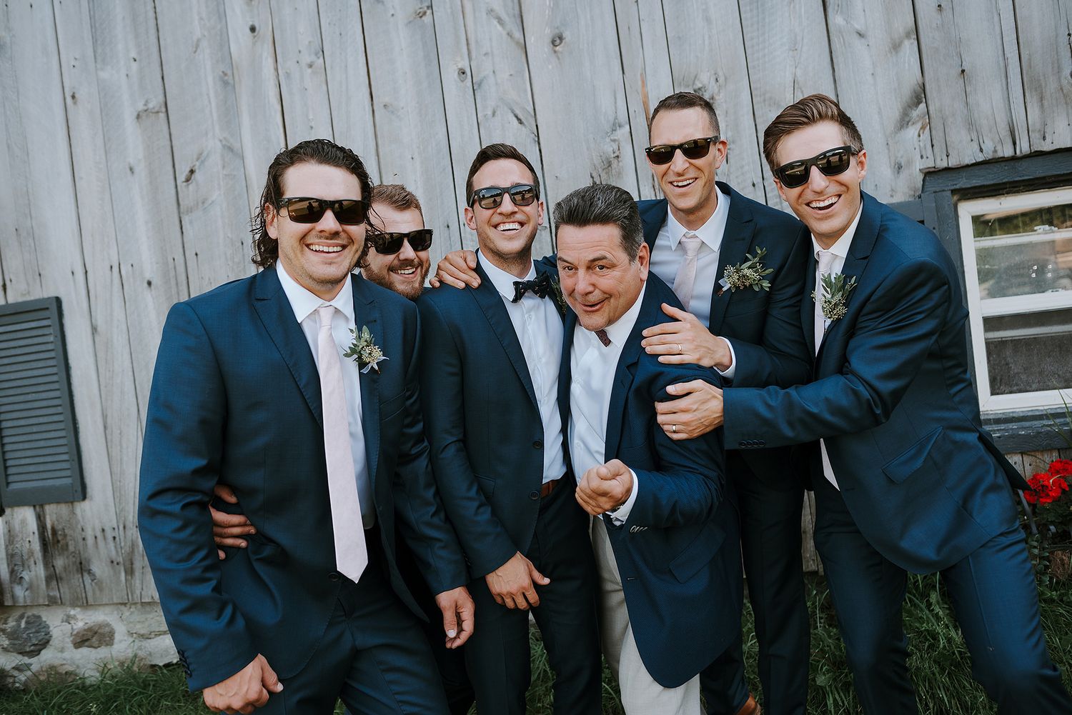 A group of groomsmen in matching navy suits and sunglasses pose together laughing against a rustic barn wall.