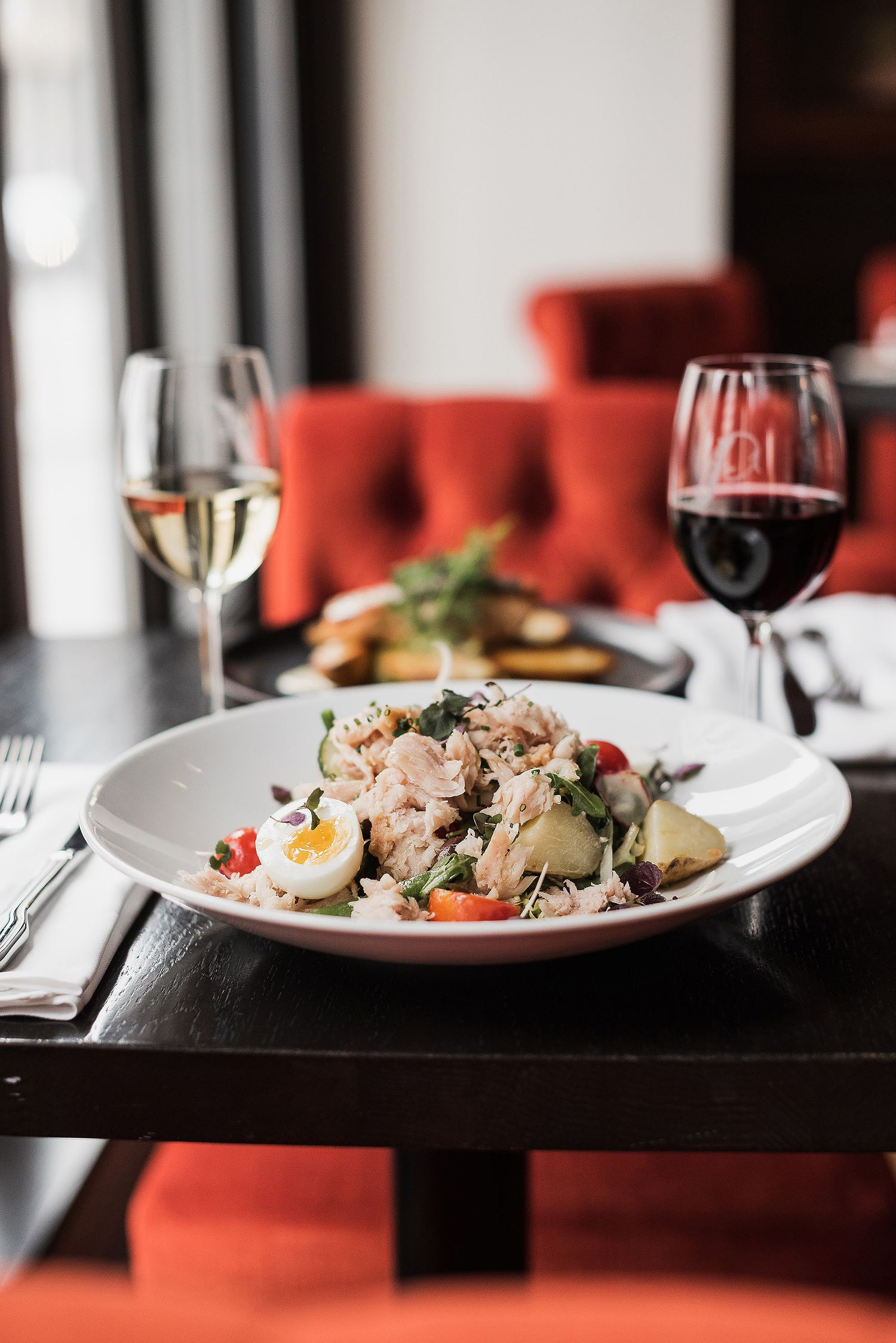 Elegant plated seafood dish with wine glasses on dark table against red chairs in upscale restaurant setting.
