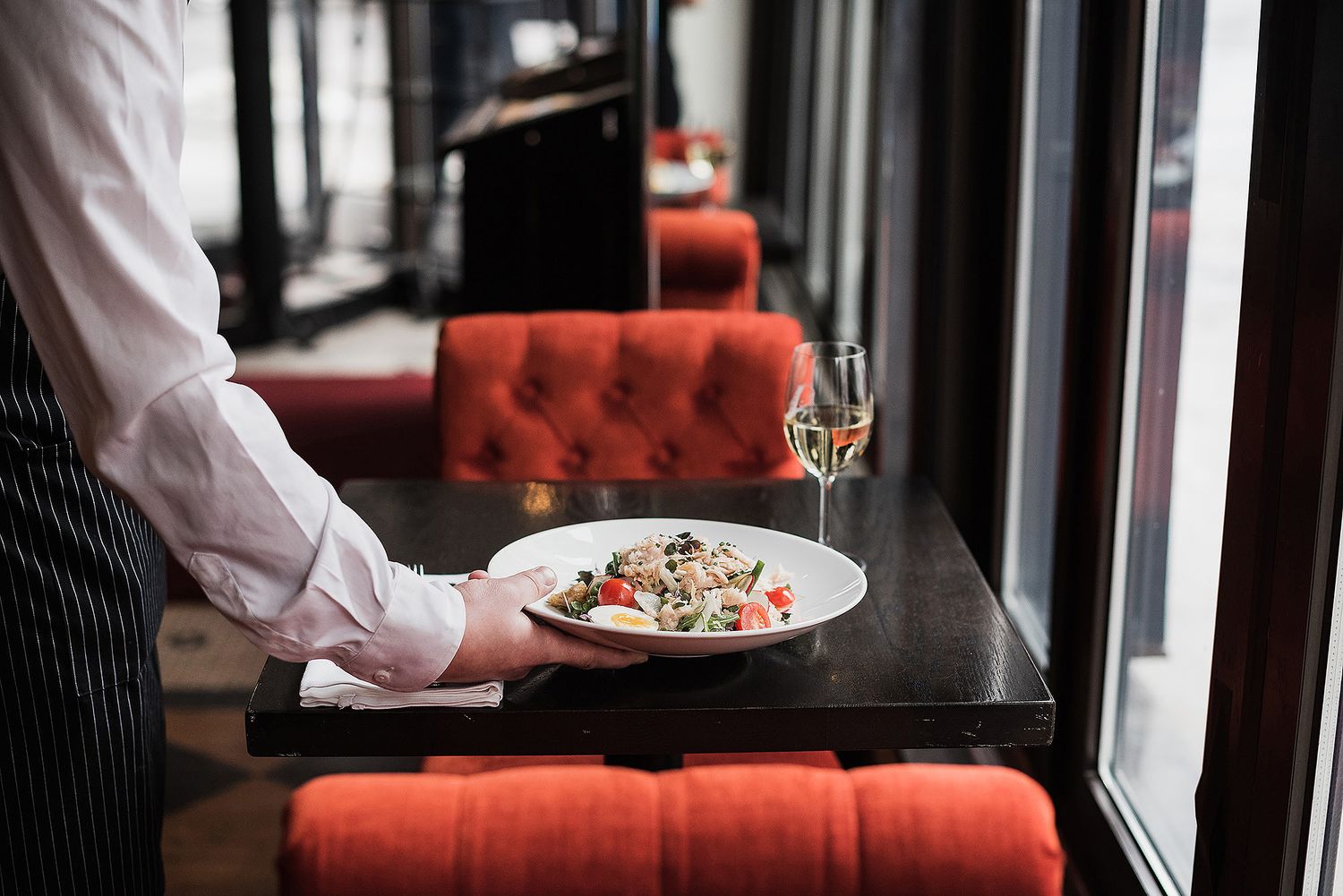 Restaurant server carrying plate of food through a modern dining room with coral-colored upholstered benches.