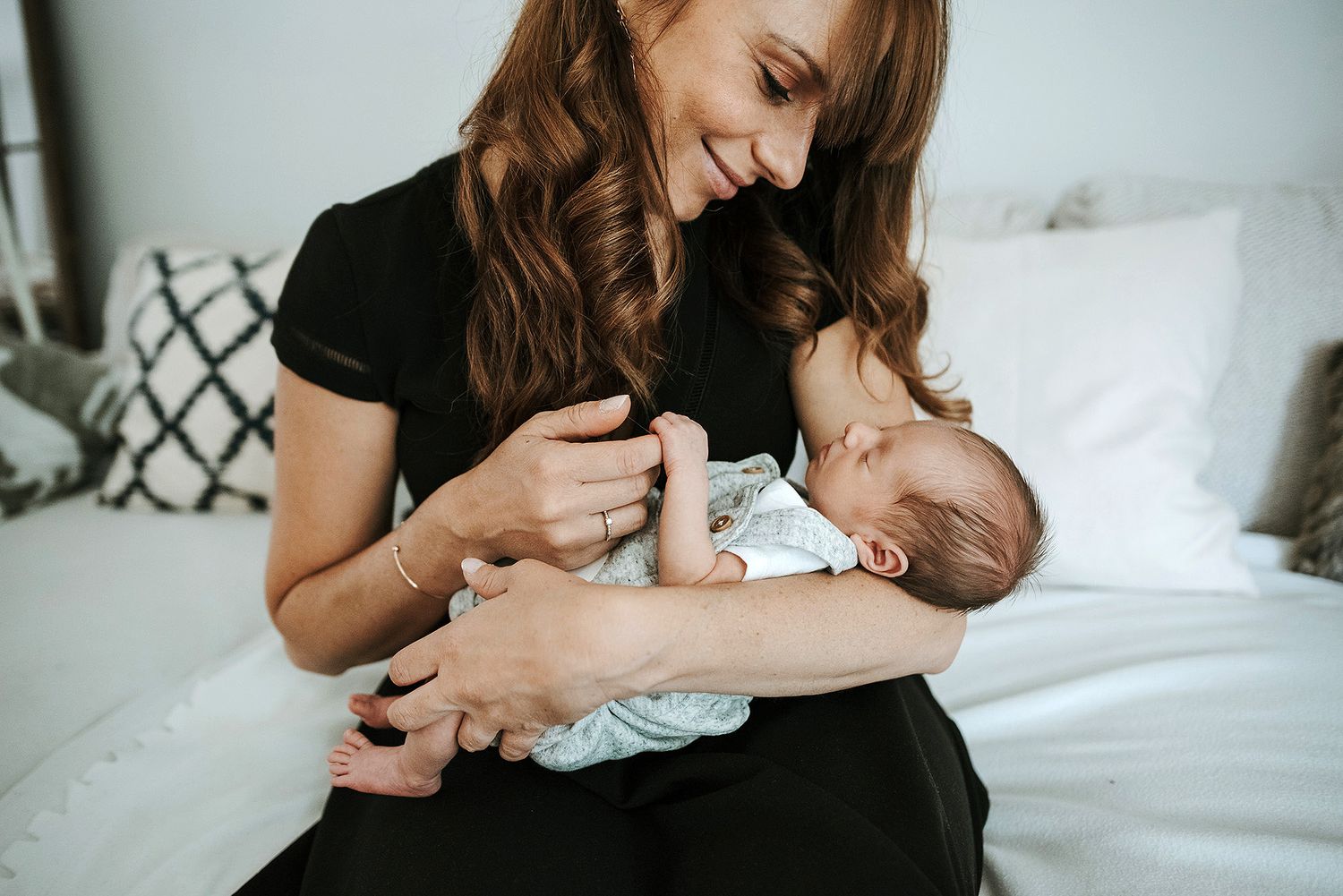 A person in a black dress sits on a bed holding and feeding a sleeping baby in white.