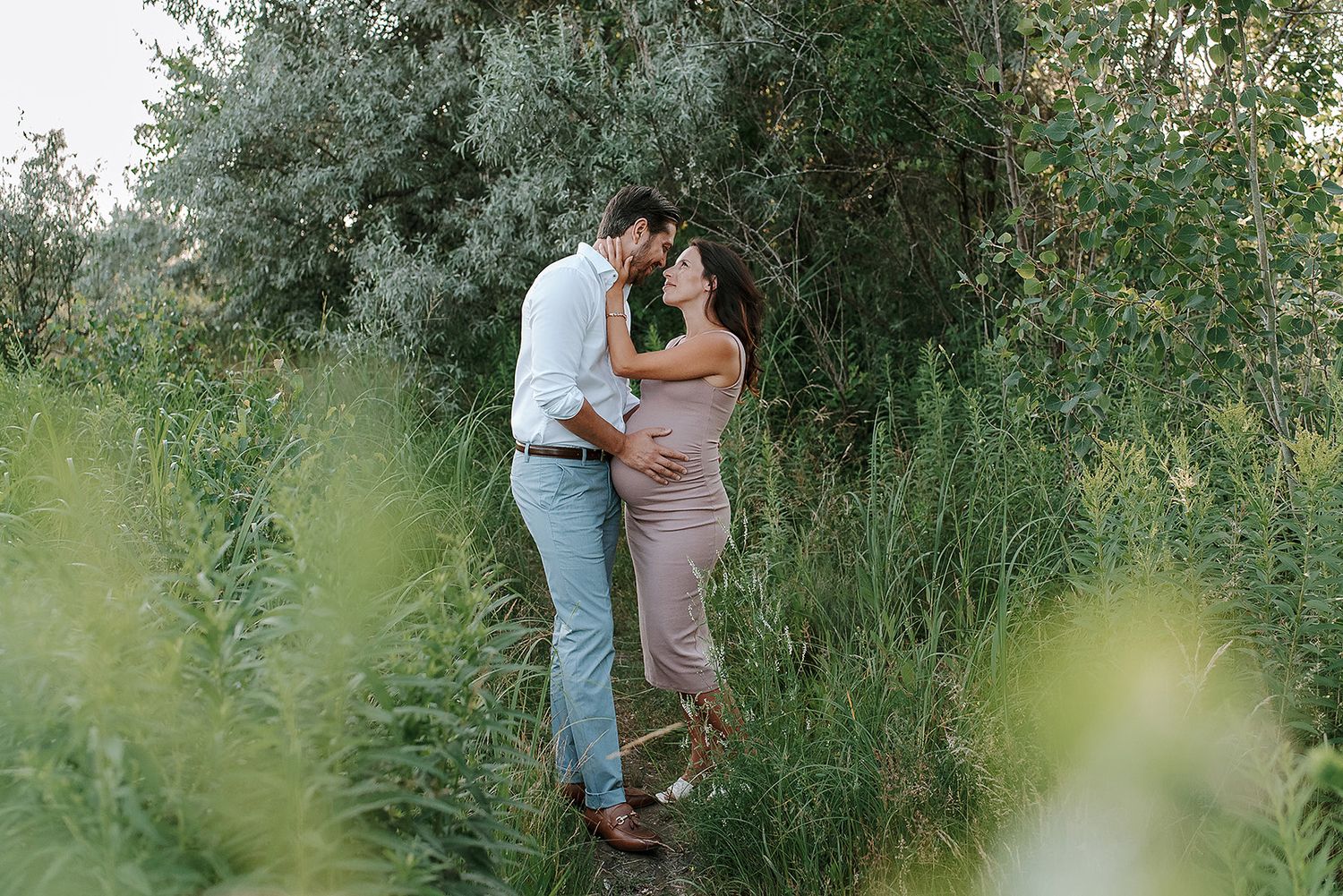Couple embraces in a romantic moment surrounded by lush green foliage and olive trees on a summer afternoon.