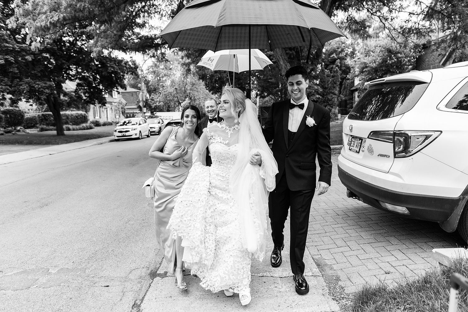 A couple in formal wedding attire walk together under a black umbrella on a tree-lined street.