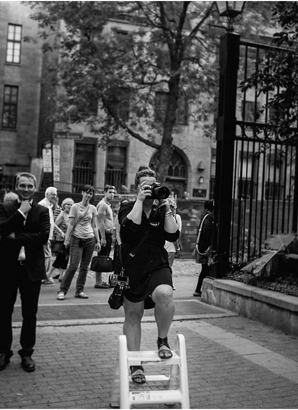 Black and white photo showing someone climbing a ladder at a campus gate while others watch on the cobblestone street.