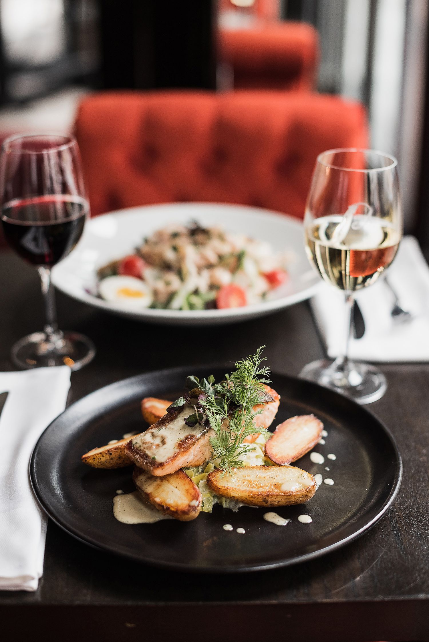 Elegant restaurant table setting with pan-seared meat, fresh herbs, and glasses of red and white wine on black plates.