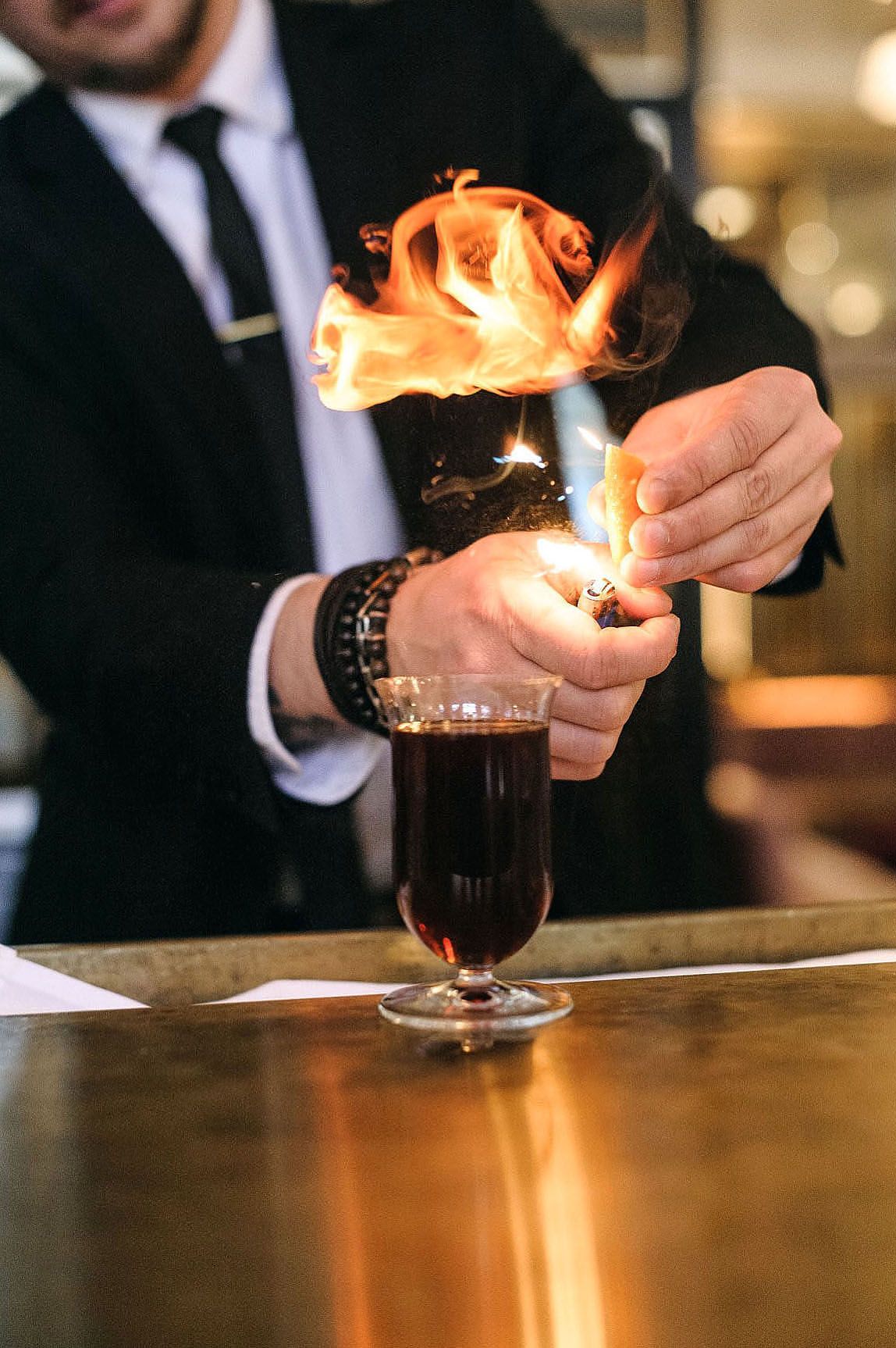 Bartender igniting flames over a dark cocktail drink in a stemmed glass on a bar counter, captured by Caroline Lessard photographe Montréal.