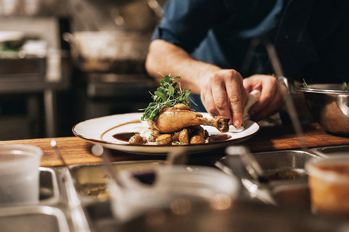 Hands plating food on a white dish with fresh herbs and roasted ingredients in a warm, moody restaurant setting.