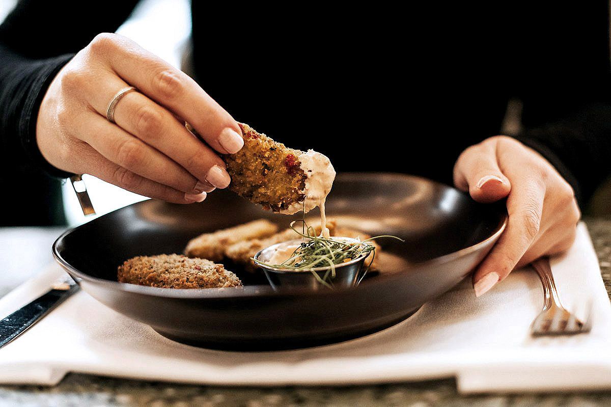 Hands holding cutlery while dining on grilled meat and herbs served on a dark ceramic plate.
