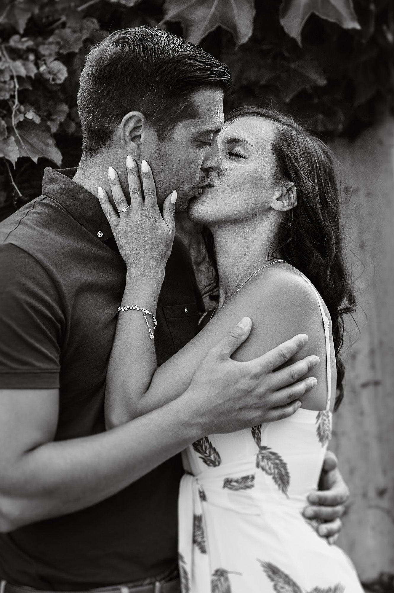 Black and white romantic portrait of a couple sharing an intimate kiss against a leafy backdrop.