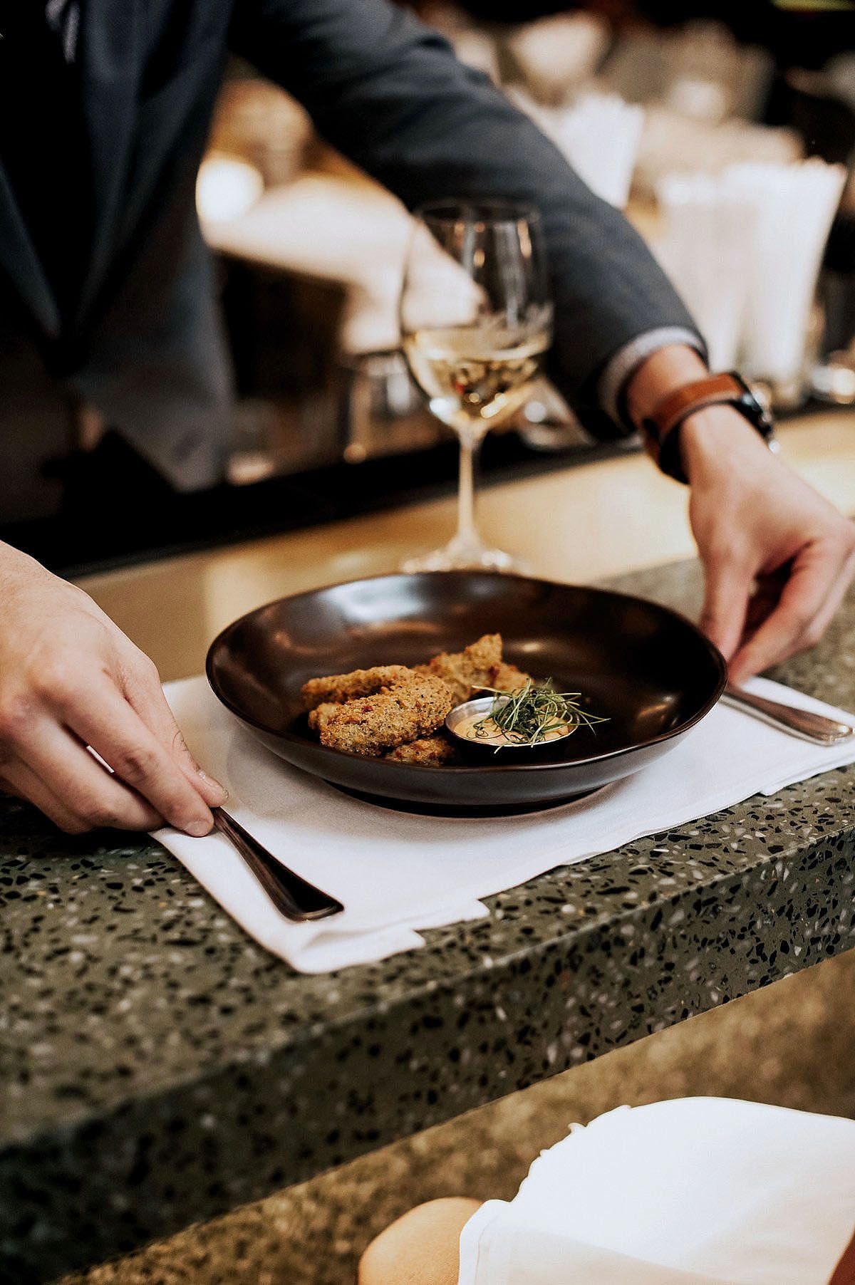 Hand serving an elegant meal with breaded meat and herbs on a dark plate, accompanied by a glass of white wine.