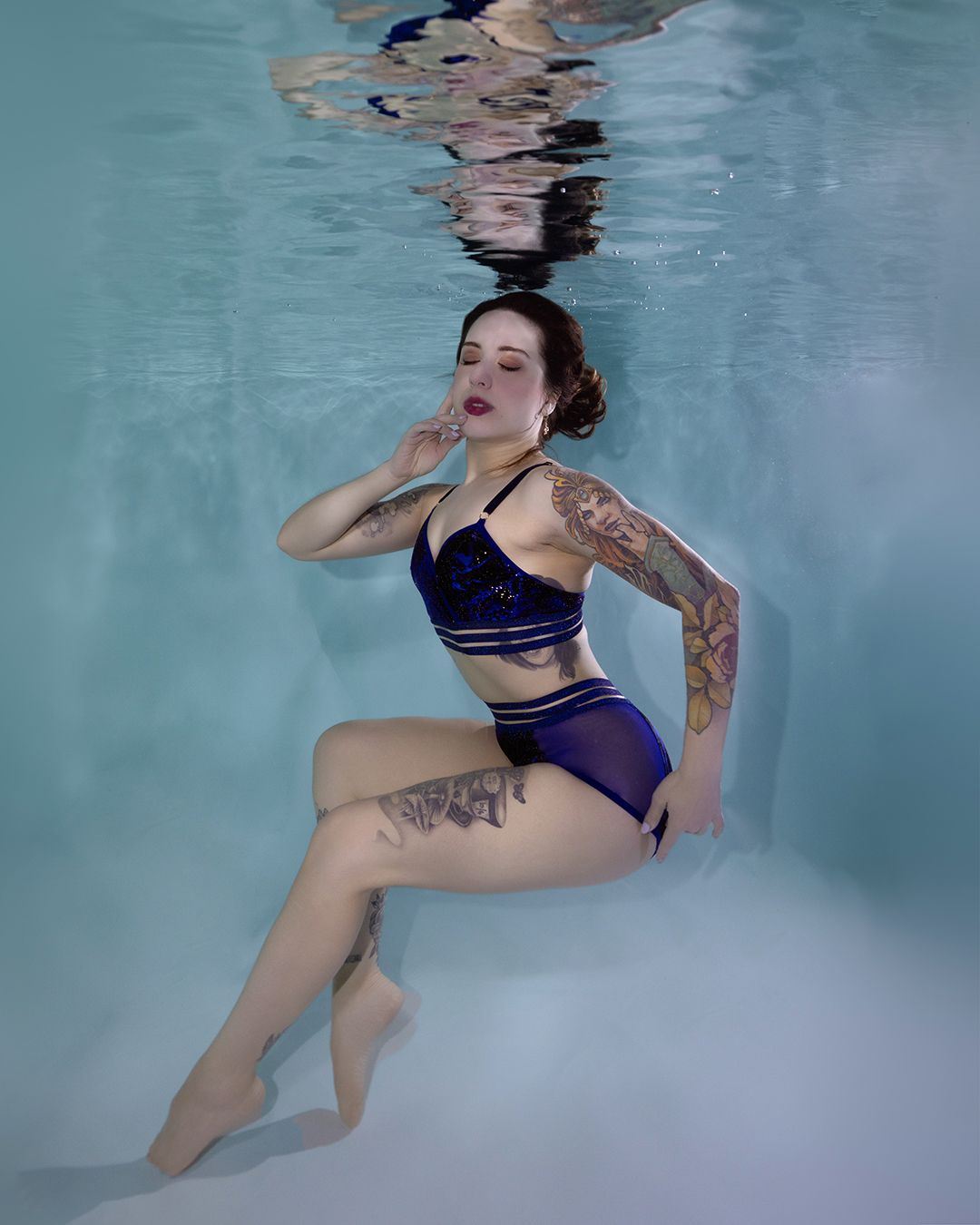Tattooed woman in blue bikini posing gracefully underwater in a pool, with light reflections on the surface above.