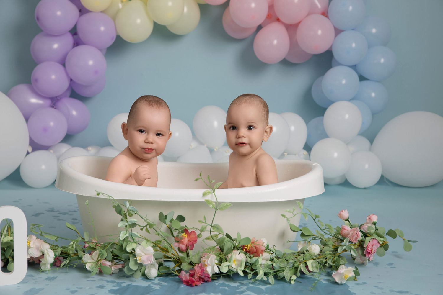 Two babies in a bathtub surrounded by pastel balloons and floral decorations for a twin photo session.