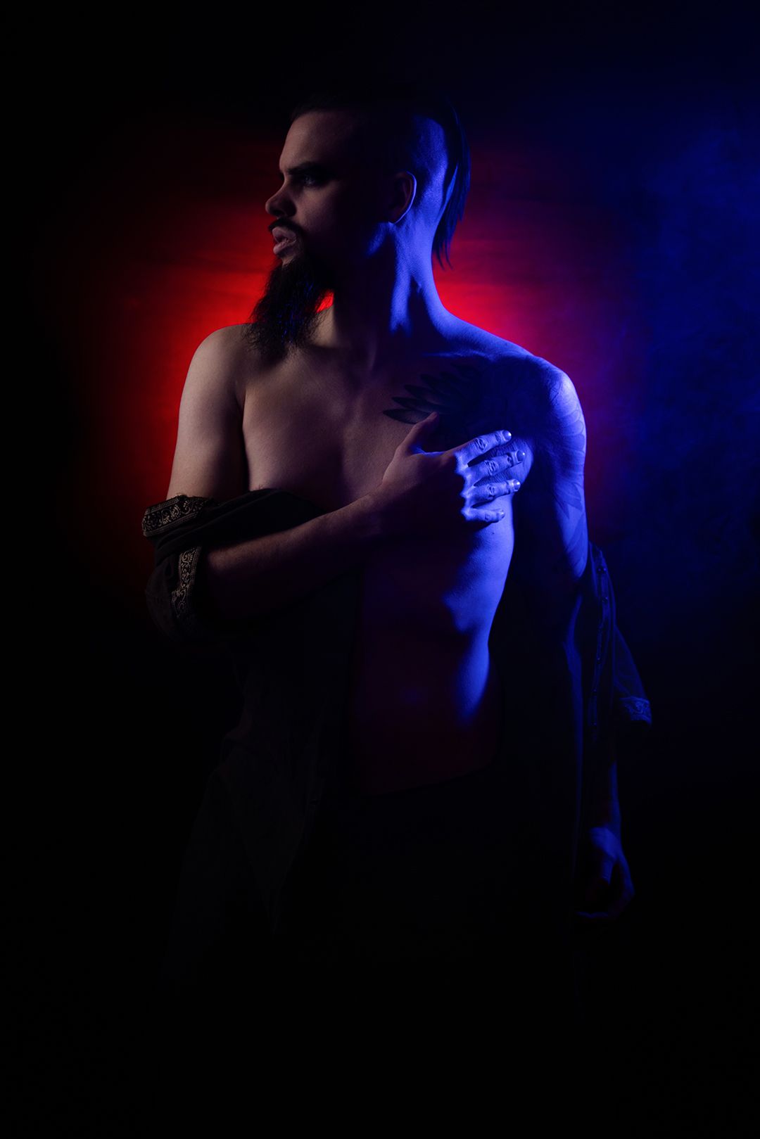 Shirtless person with long hair posed dramatically under blue and red studio lighting against a dark background.