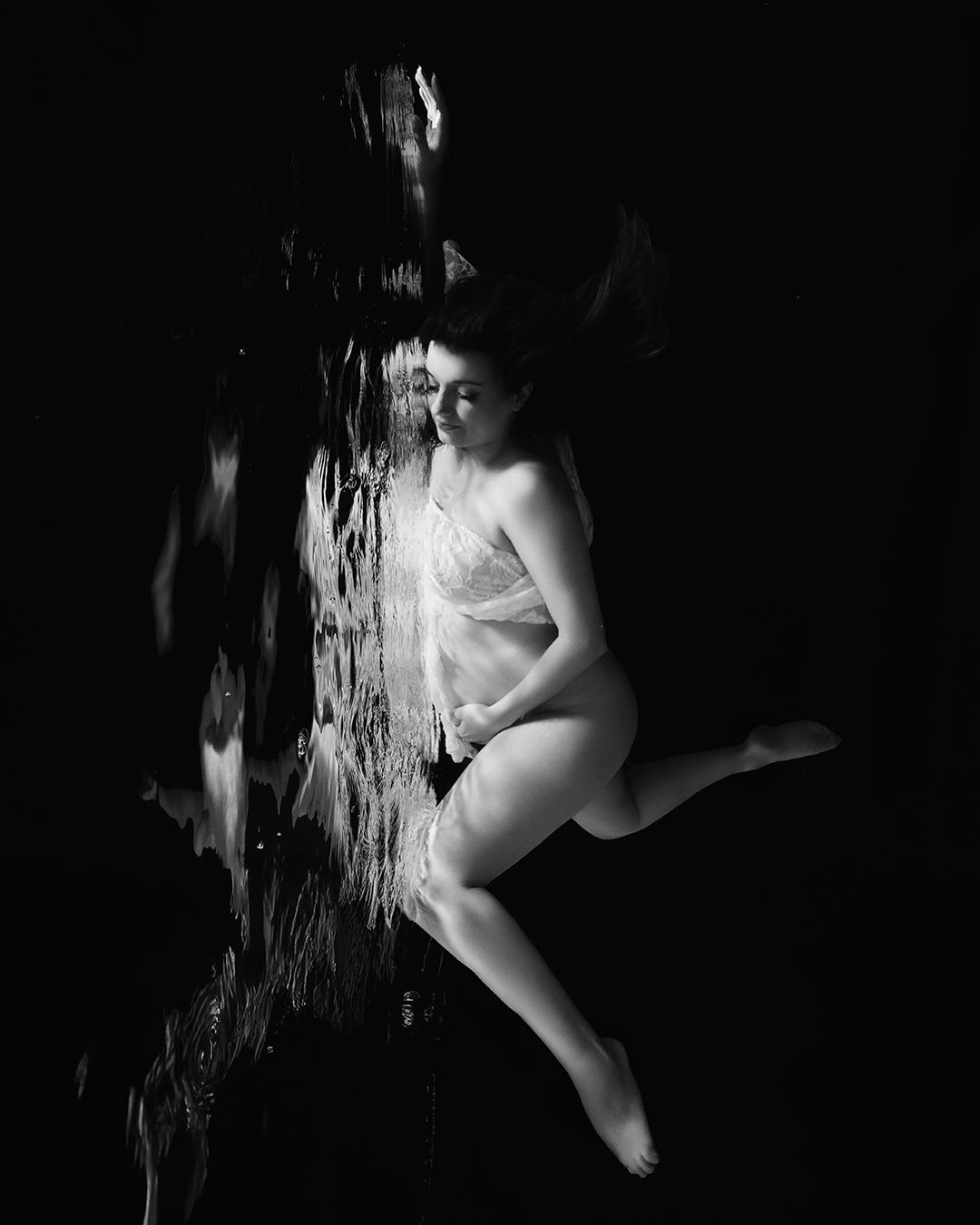 Black and white underwater photo of a woman in lingerie gracefully posed against dark water with light reflections.