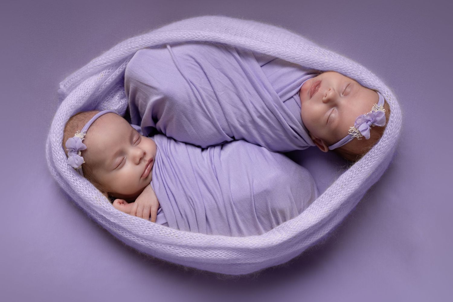 Two sleeping newborn babies wrapped in soft purple fabric lie nestled together in a cozy oval basket.