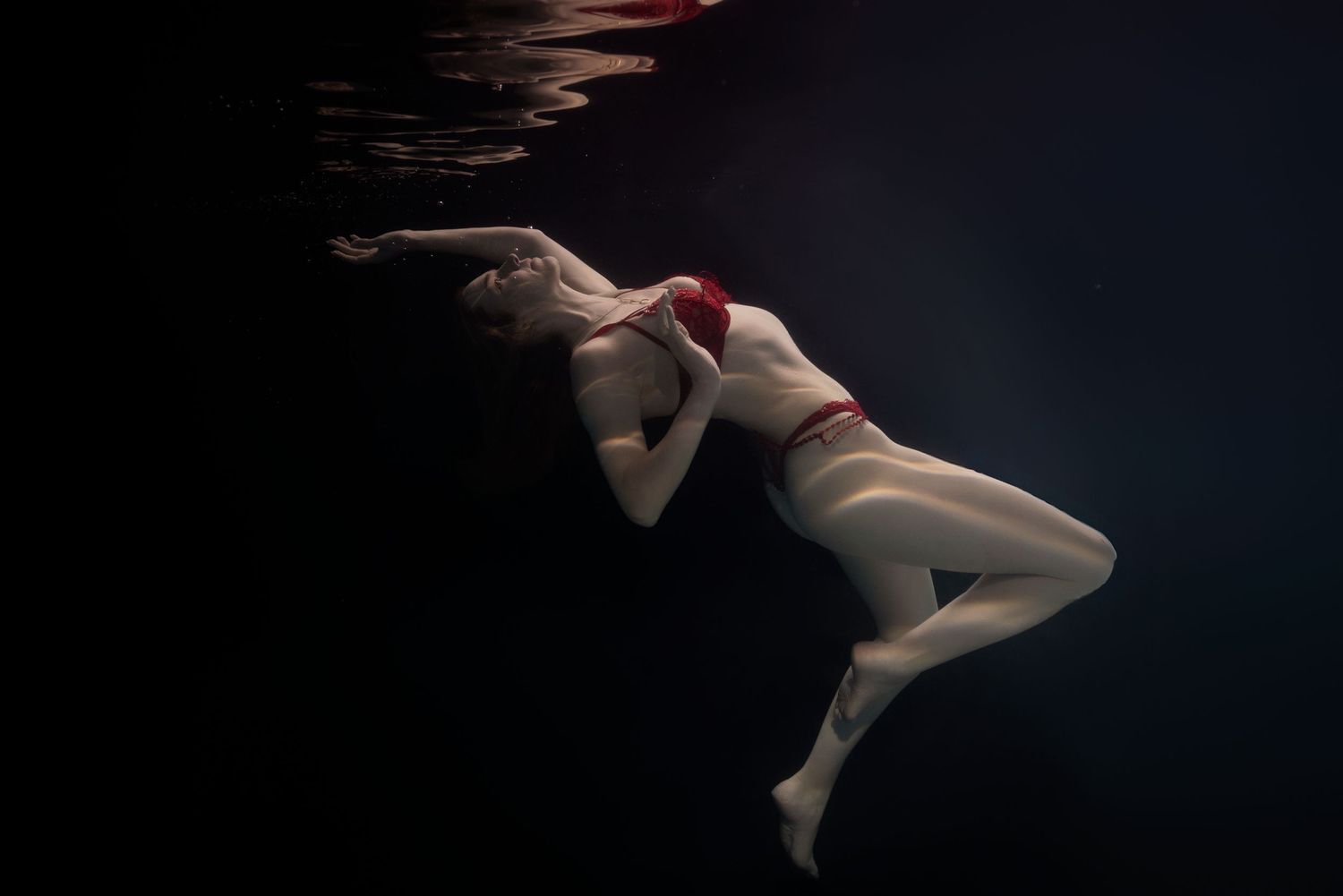A woman in a red bikini top and white leggings floats gracefully underwater against a dark background.