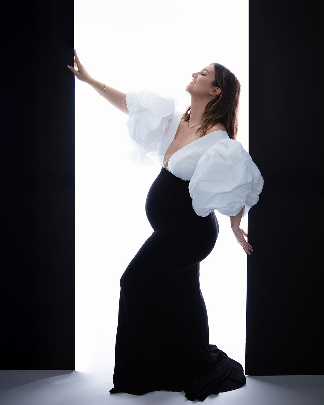 A pregnant woman in a black dress and white sleeves poses gracefully against a white backdrop in an artistic maternity photo.