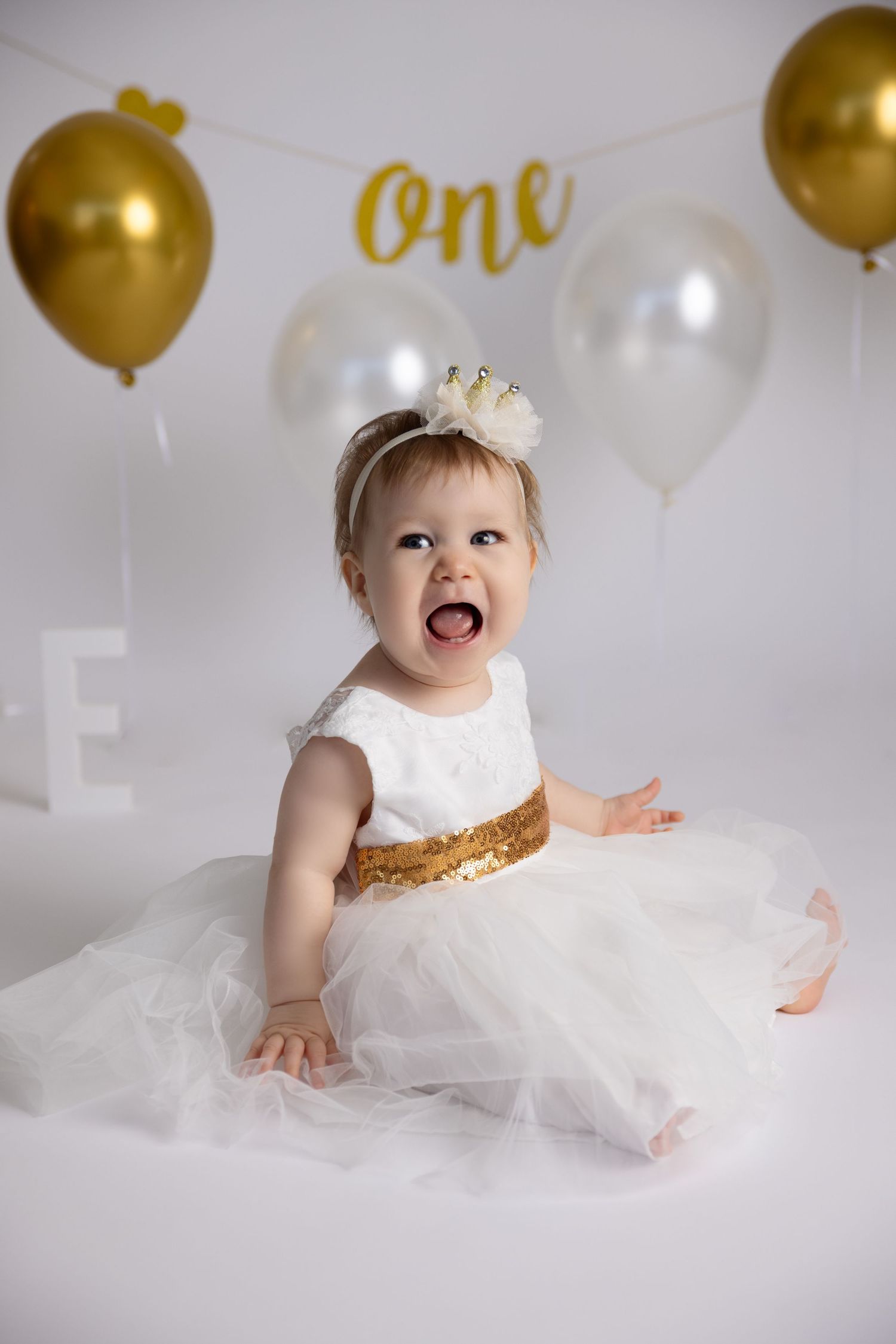 A baby in a white tulle dress and gold crown poses with gold balloons for a first birthday photoshoot.