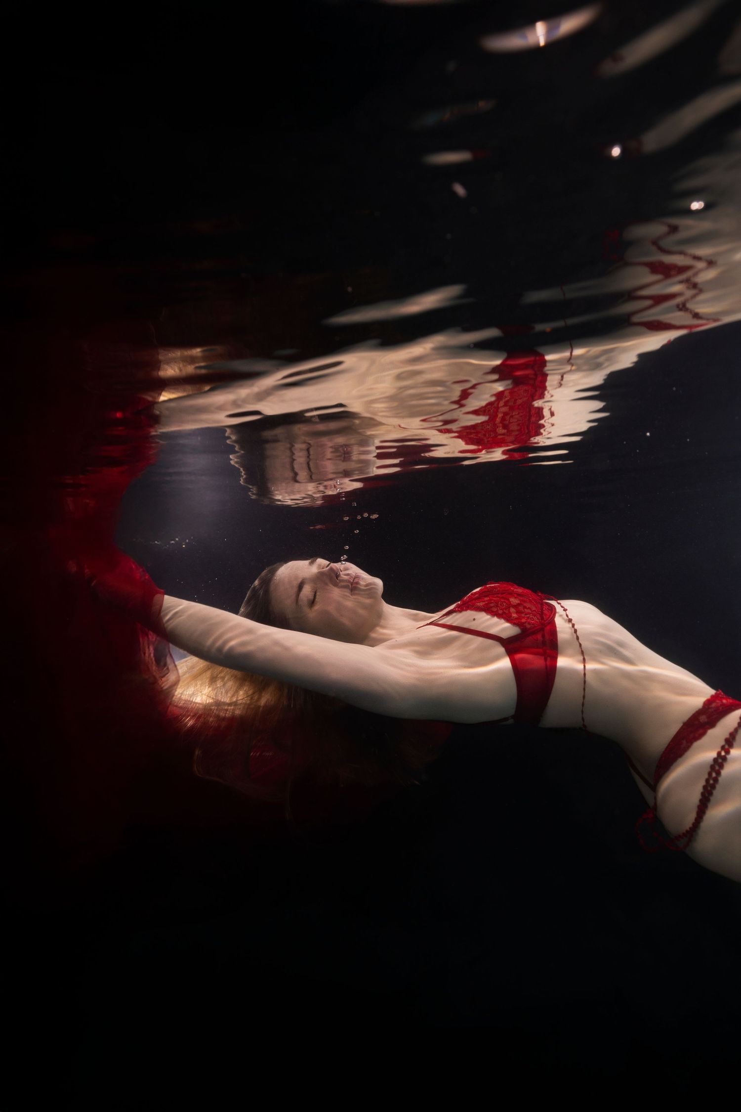 Woman in red swimsuit floating underwater in dark pool, viewed from below with light reflections on surface.