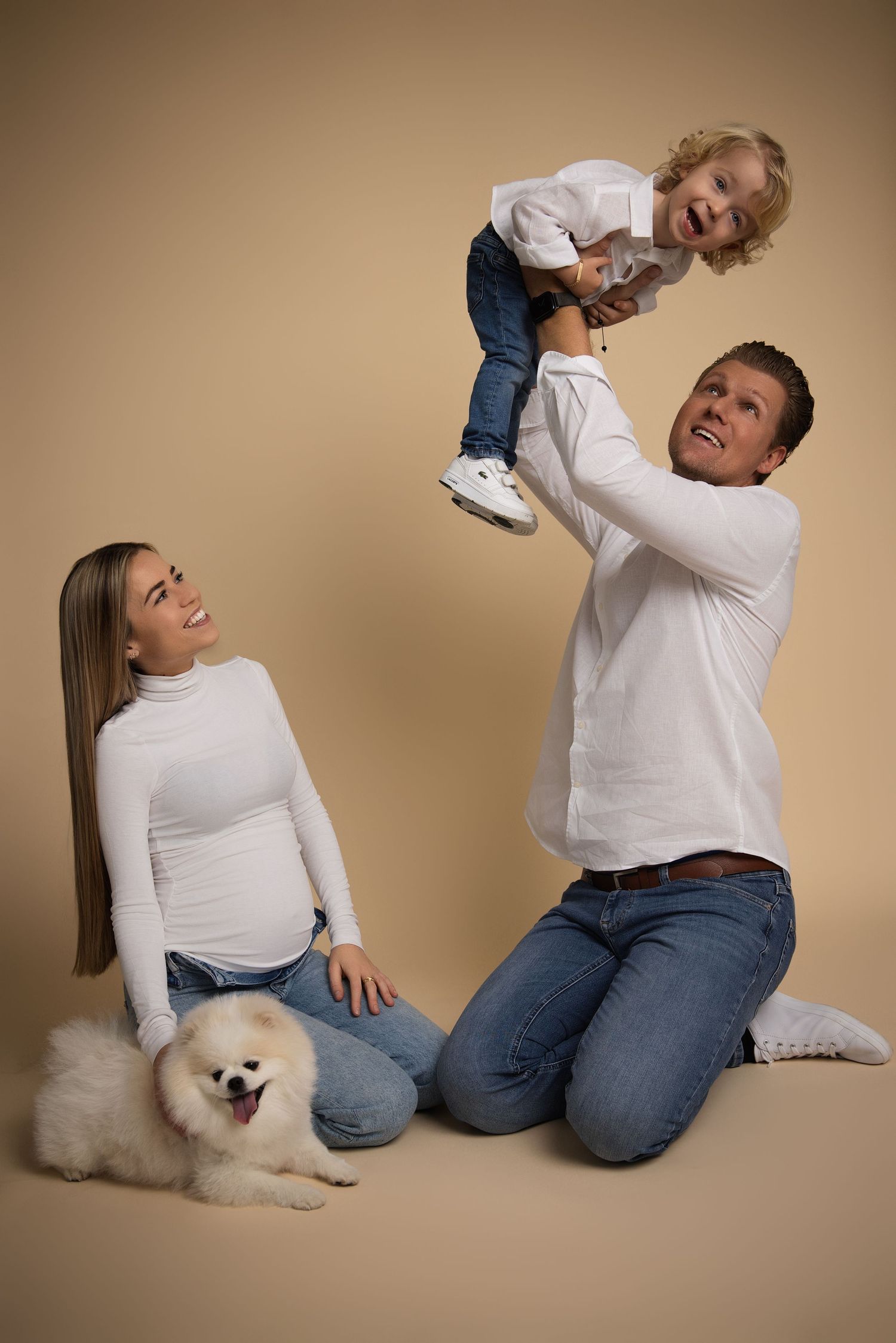 Playful family portrait with a white fluffy Pomeranian dog on a beige studio background.