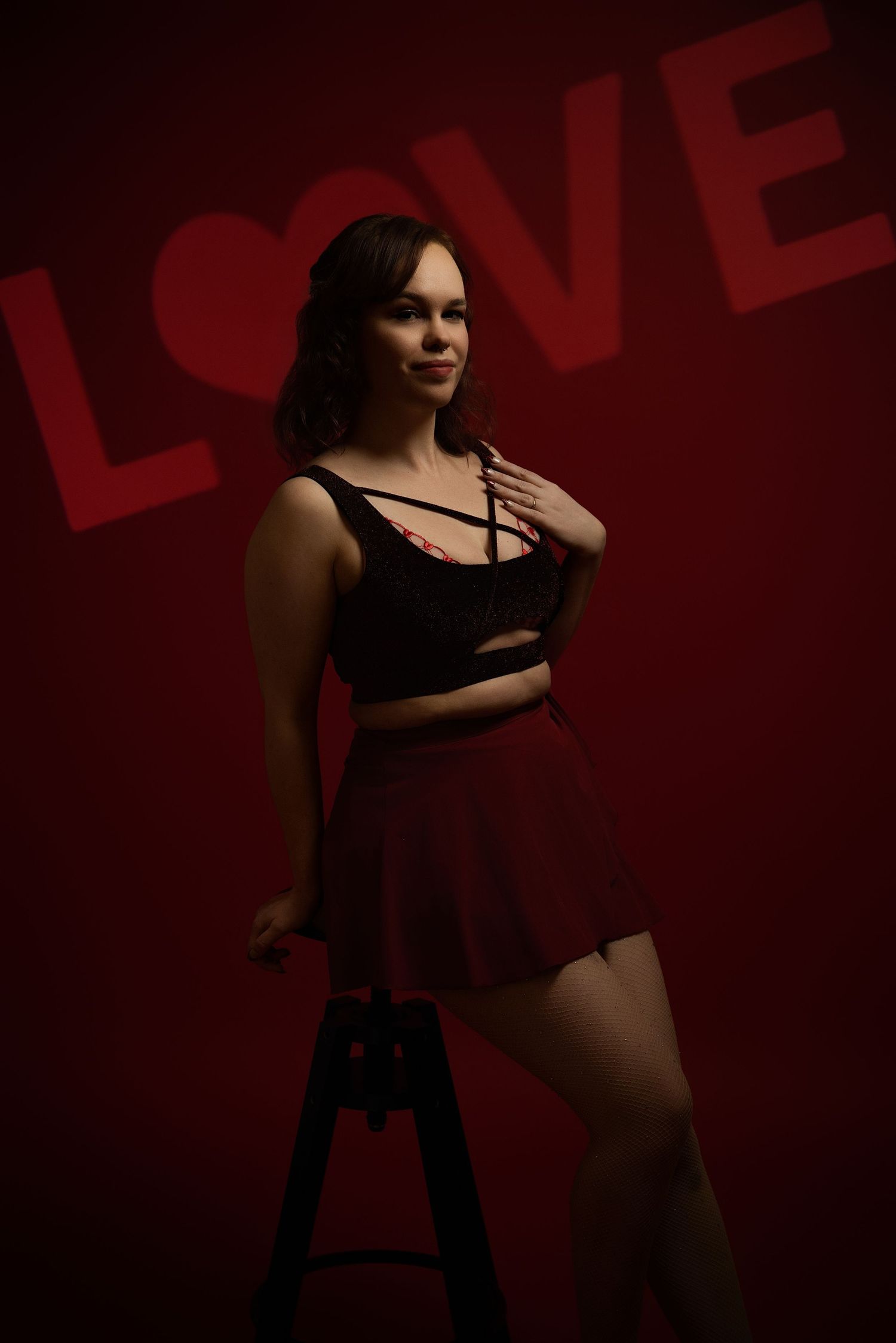 Young woman in black crop top and red skirt poses confidently against a dark red background with LOVE text.