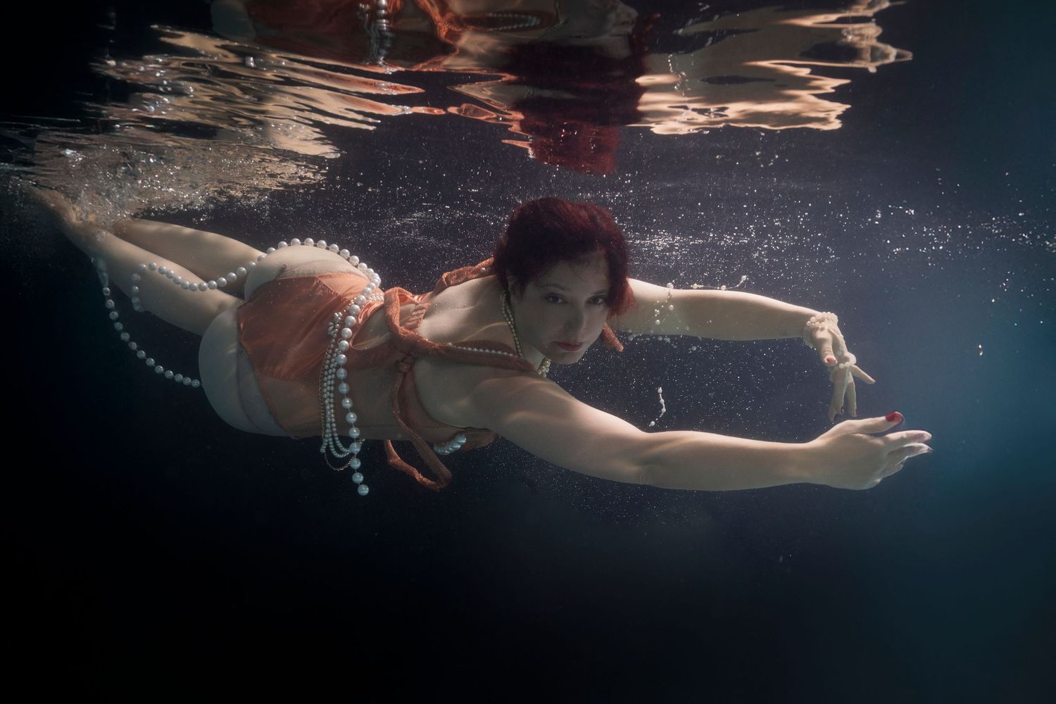 Woman swimming underwater in a red outfit adorned with pearl necklaces, gracefully gliding through dark water.