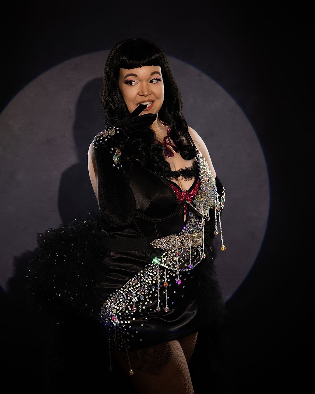 A performer in a black mini dress adorned with sparkling rhinestones and a feather boa poses under a spotlight.