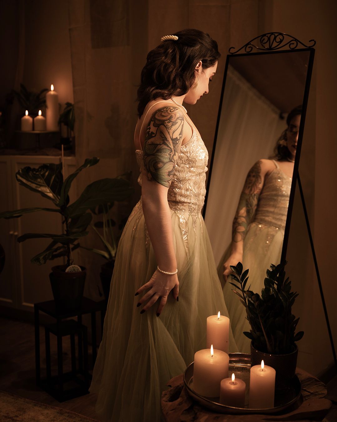 Woman in elegant sage green gown stands before a mirror surrounded by glowing candles in a warmly lit room.
