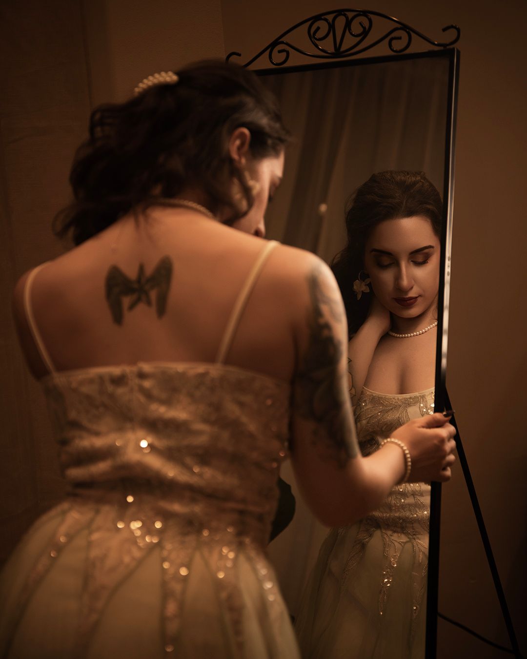 Tattooed woman in sparkly white gown gazes at her reflection in an ornate standing mirror in warm light.