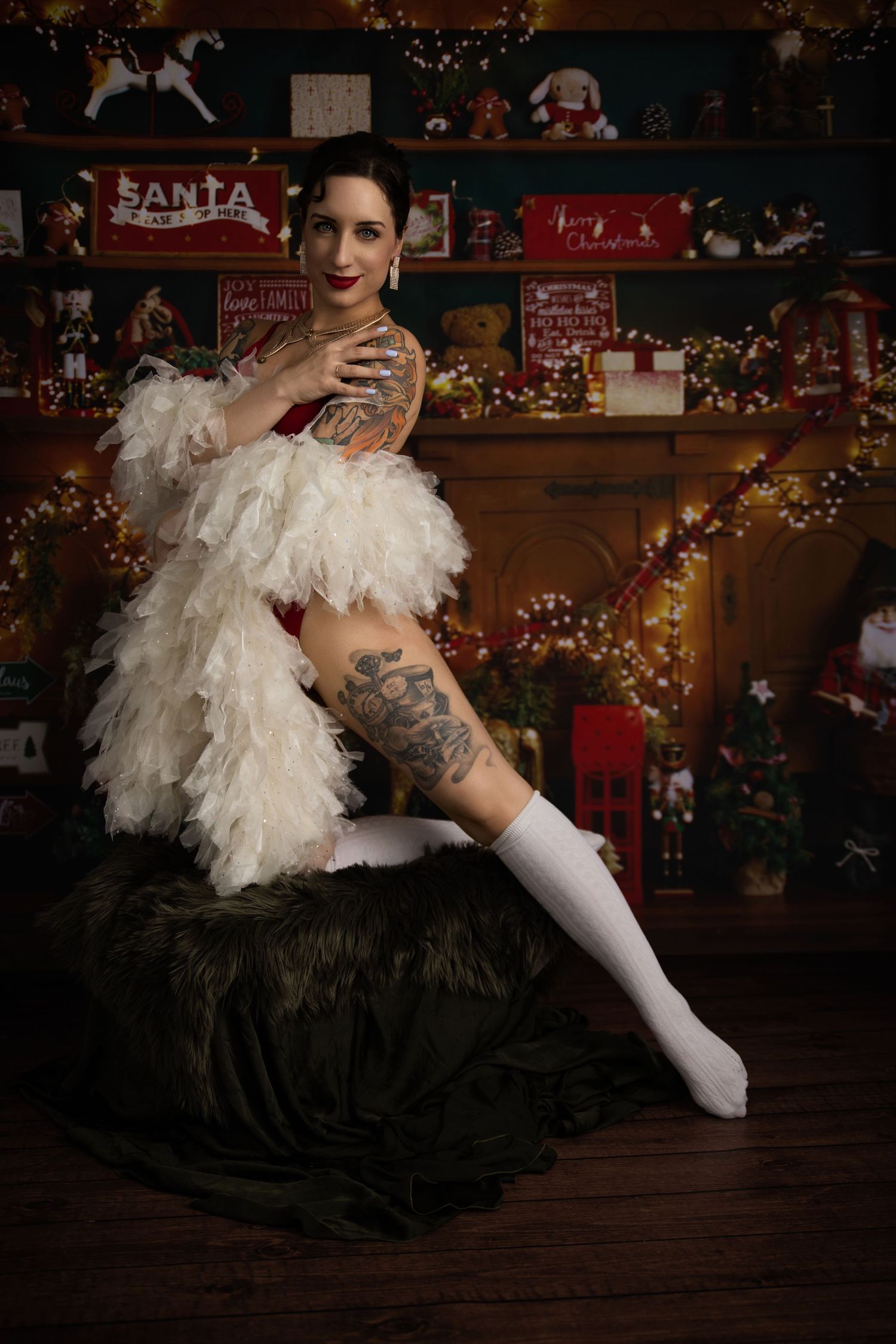 Pin-up style boudoir photoshoot in festive holiday setting with white stockings and feathers against dark backdrop.