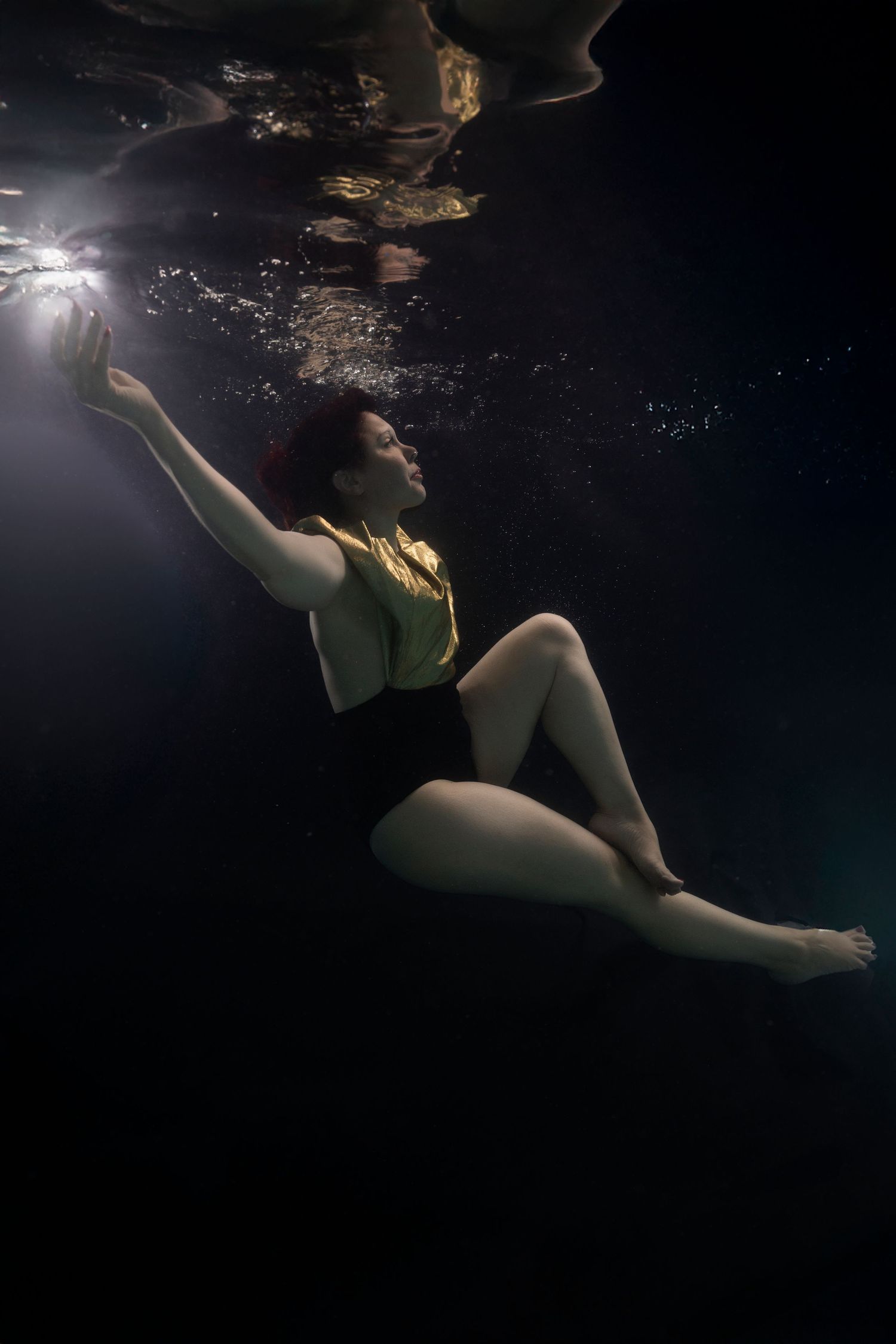 Woman in black swimsuit reaching upward underwater in dramatic dark pool with light rays streaming down.