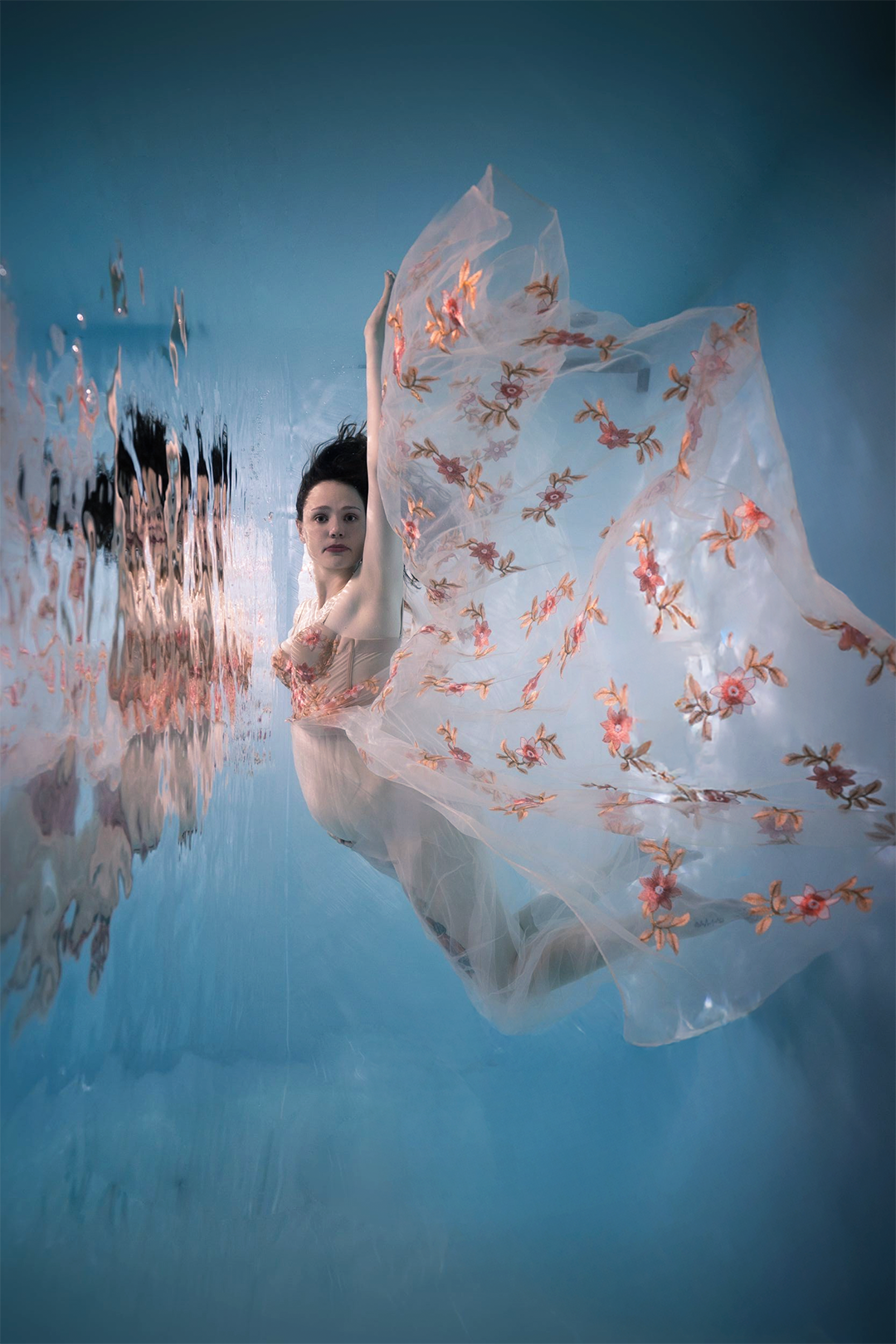Woman in floral embroidered sheer gown floating underwater in a dreamy blue aquatic setting.