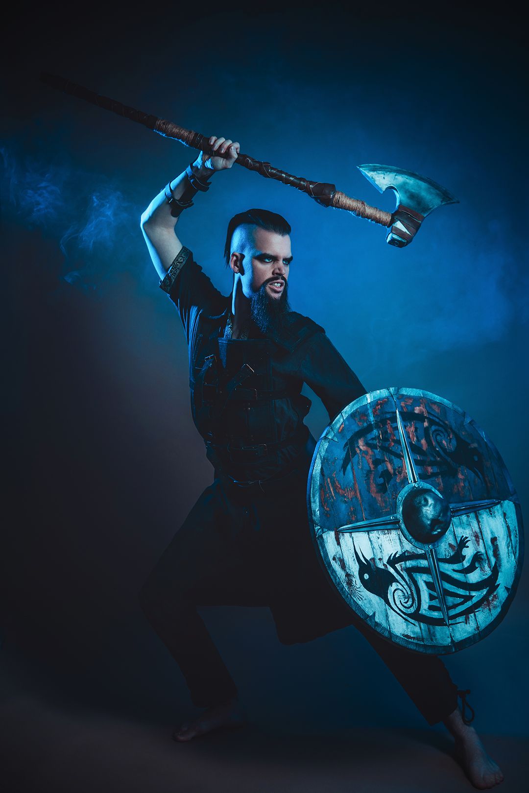 Fierce Viking warrior in black wielding an axe and decorated shield in dramatic blue-lit studio setting.