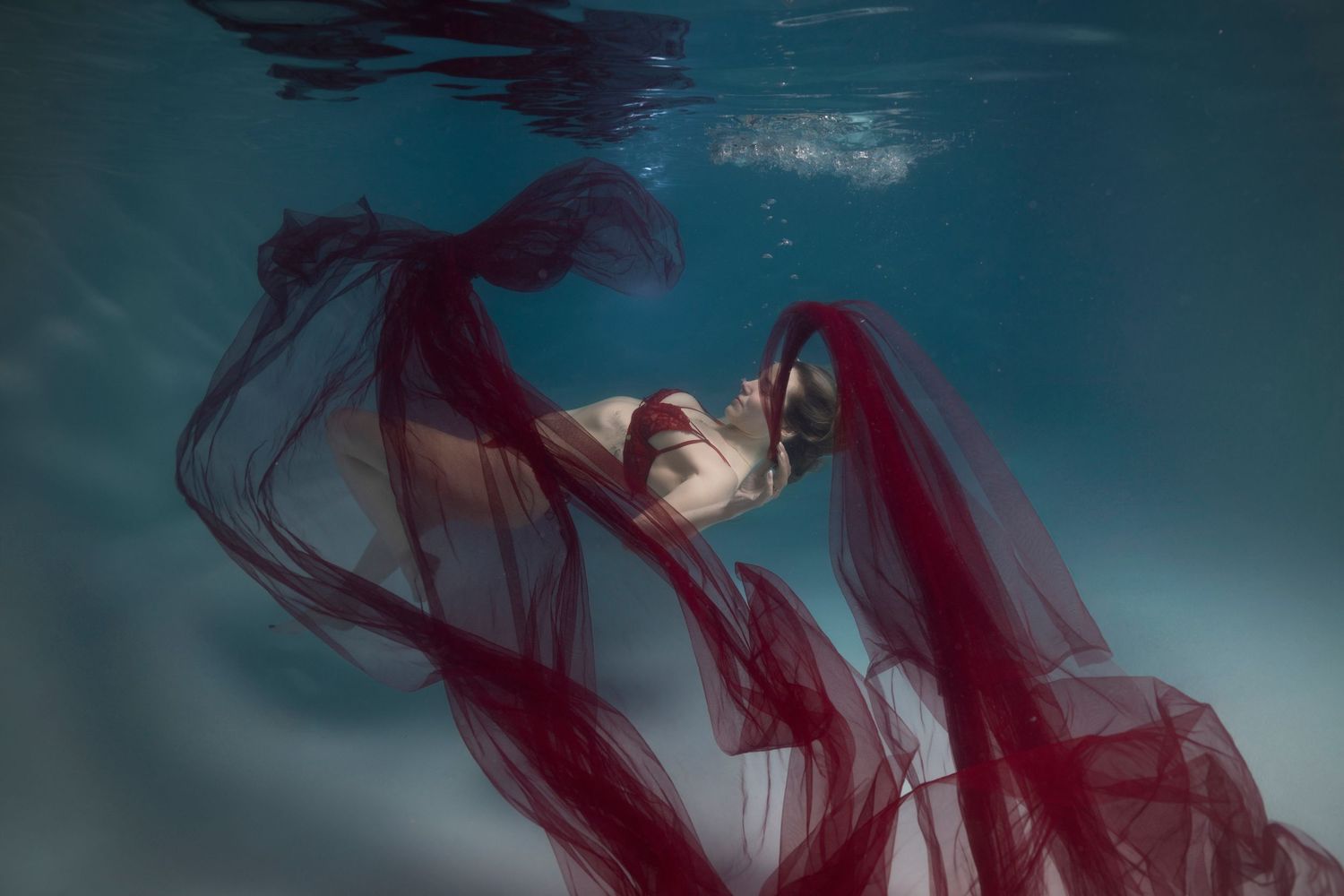 A woman draped in flowing red fabric poses gracefully underwater in a serene blue pool.