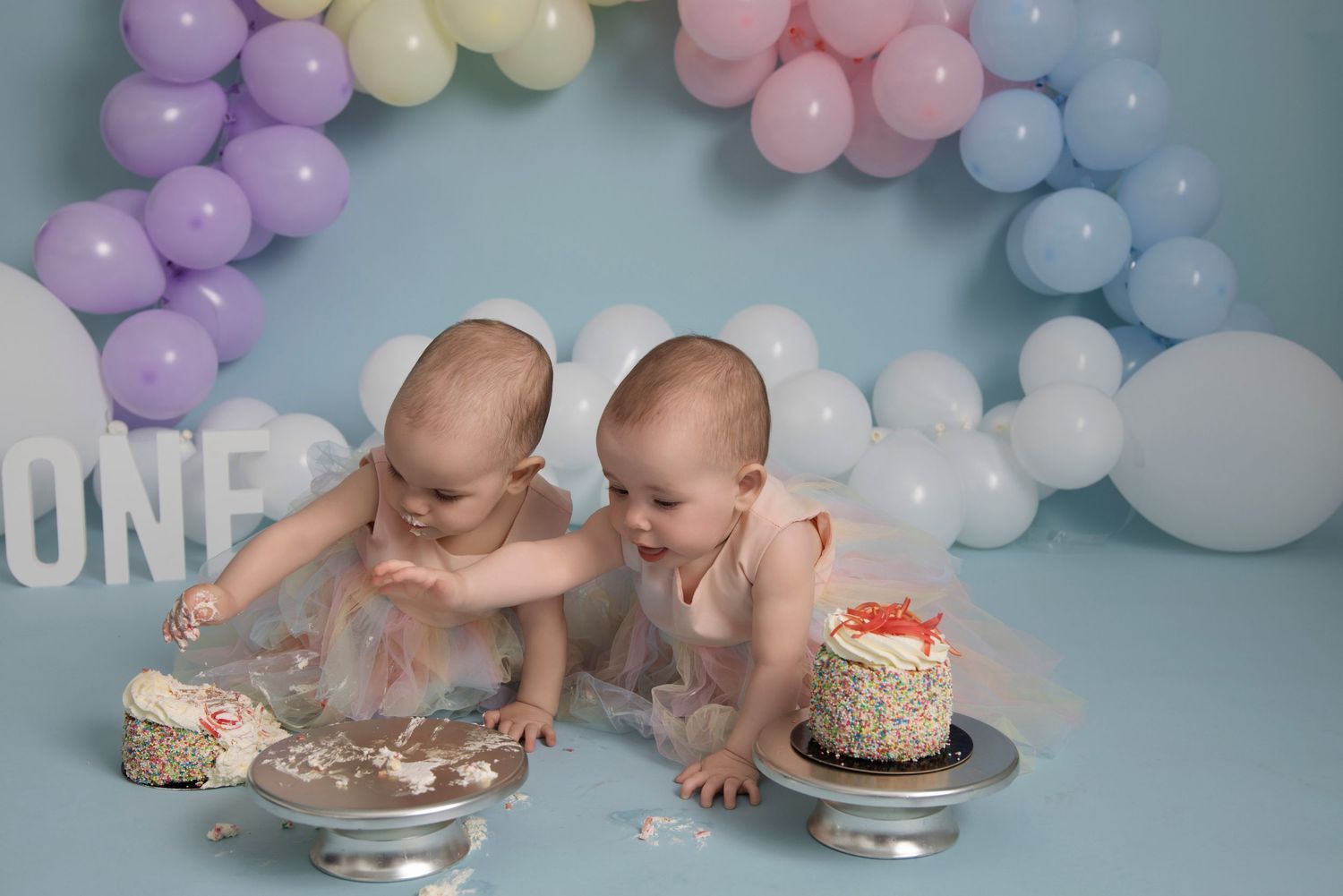Two babies enjoying a cake smash photo session with pastel balloon arch backdrop and silver cake plates on blue floor.