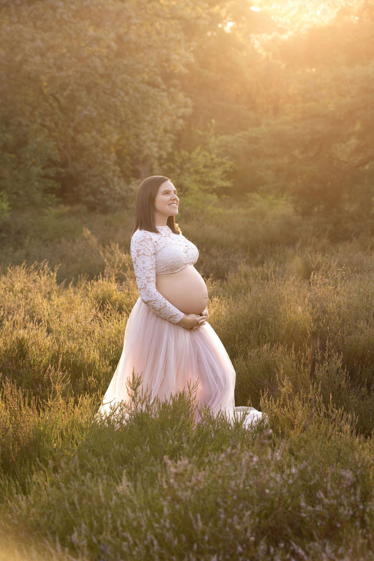 A dreamy maternity photo session in a golden wildflower field at sunset shows a woman in a flowing white lace dress.