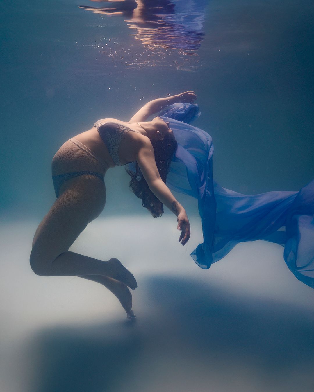 Pregnant woman floating underwater in bikini with flowing blue fabric, captured in a serene maternity photo session.