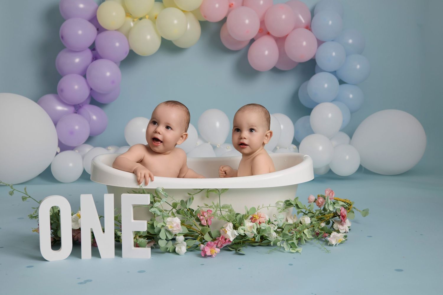 Babies sit in a decorated bathtub under a pastel balloon arch with floral decorations and 'ONE' text display for first birthday.