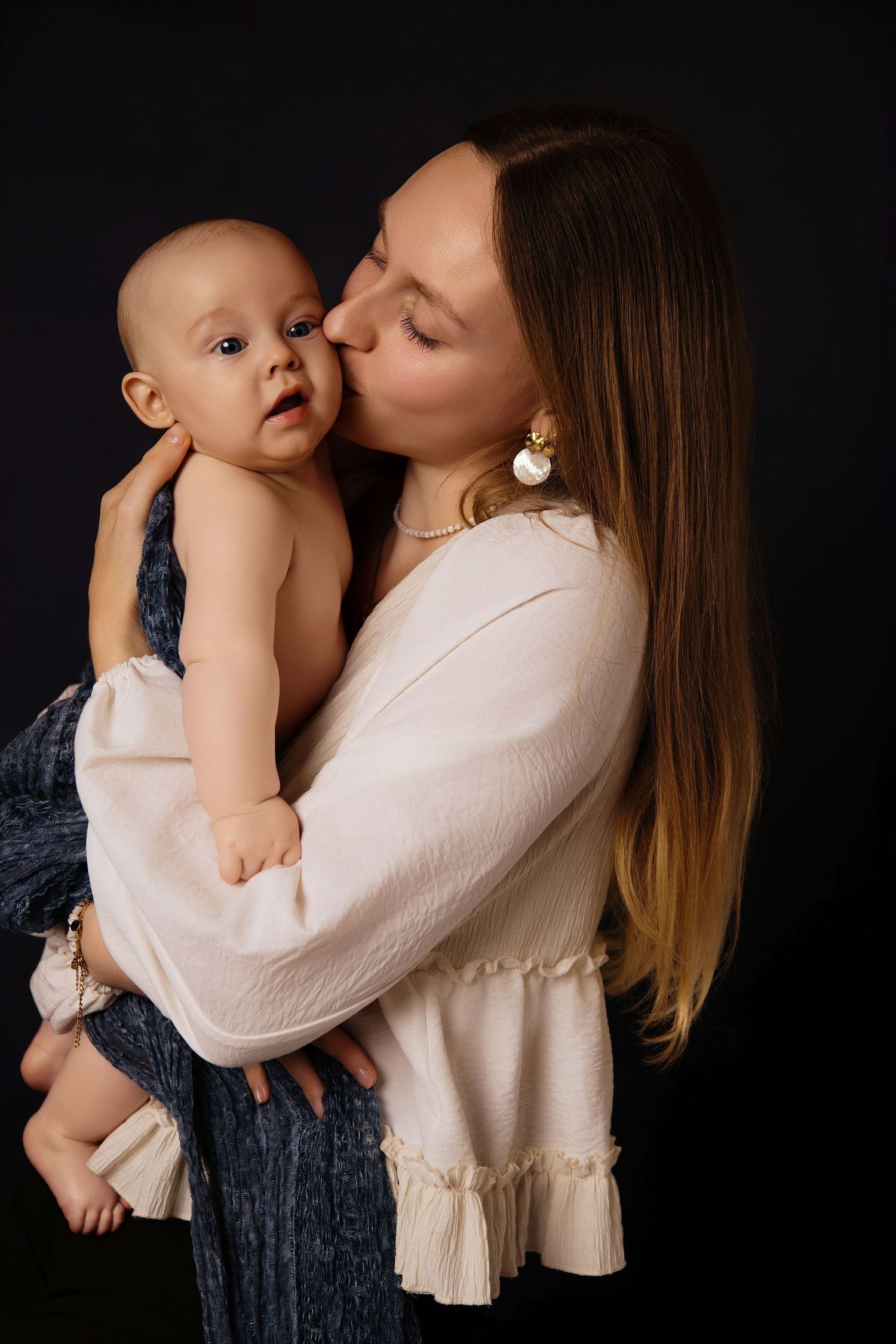 A tender moment captured against a dark background as a mother in a cream blouse kisses her baby wearing navy blue.