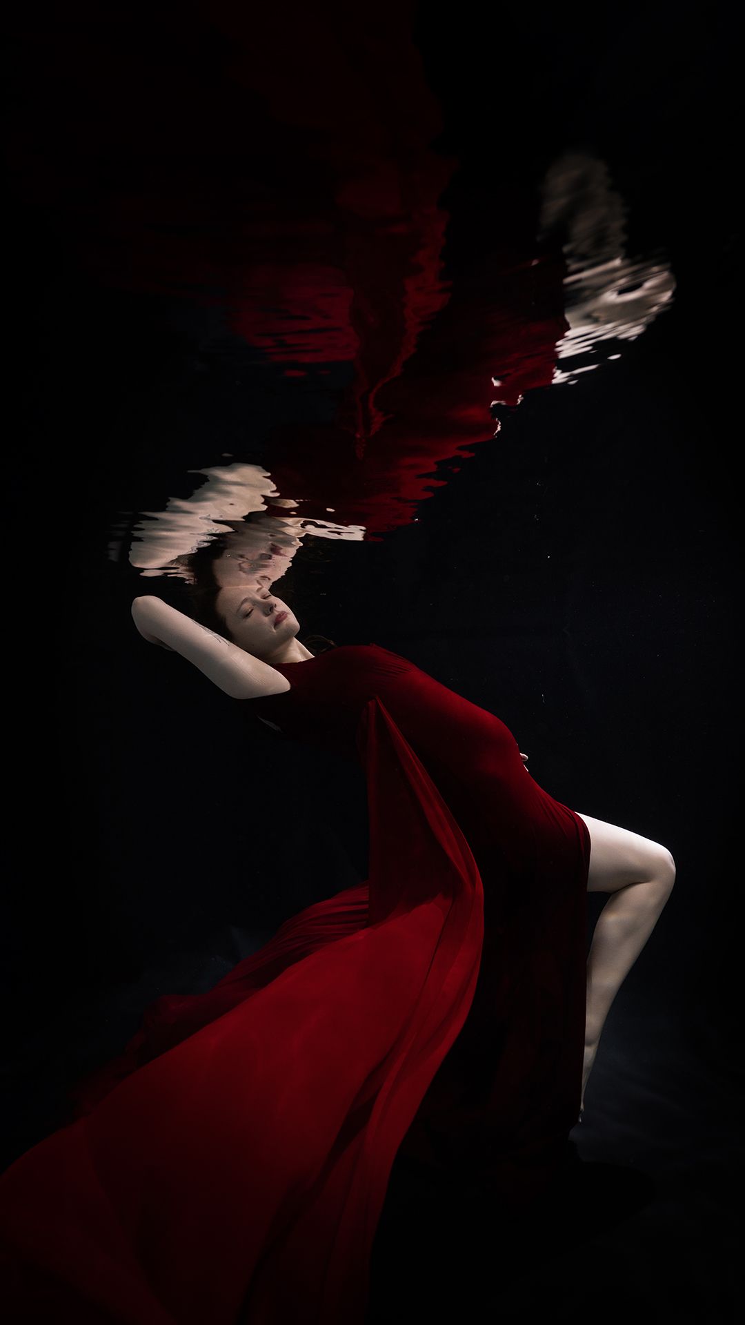 Woman in flowing red dress submerged underwater against dark background, creating dramatic artistic photography.