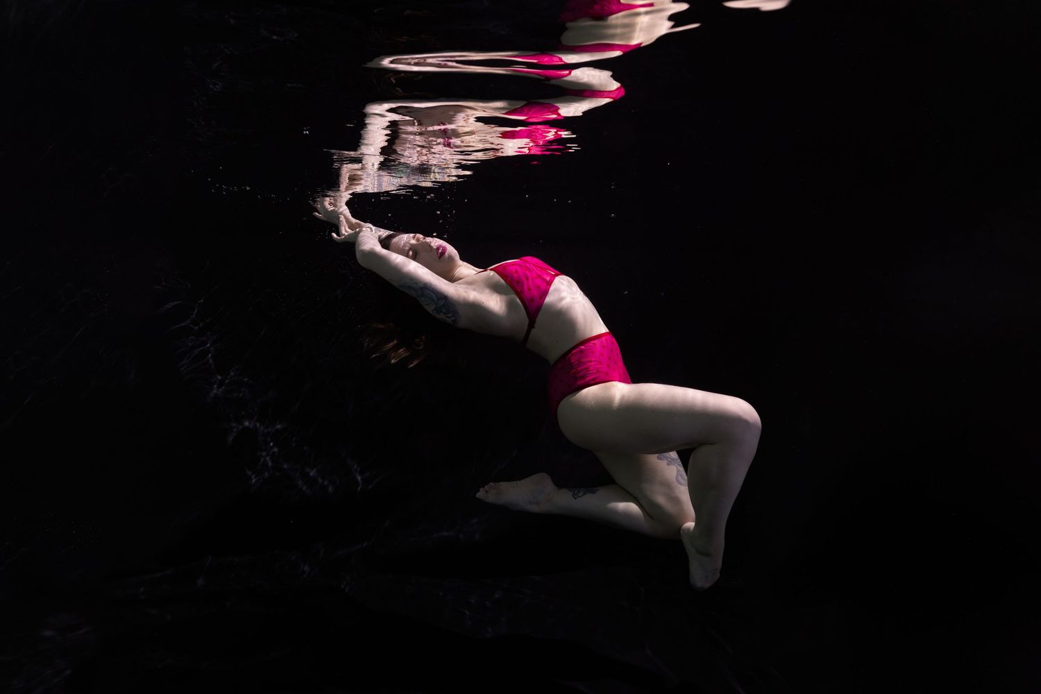 Woman in pink bikini swimming underwater against a dark background, captured in an artistic underwater photography shot.