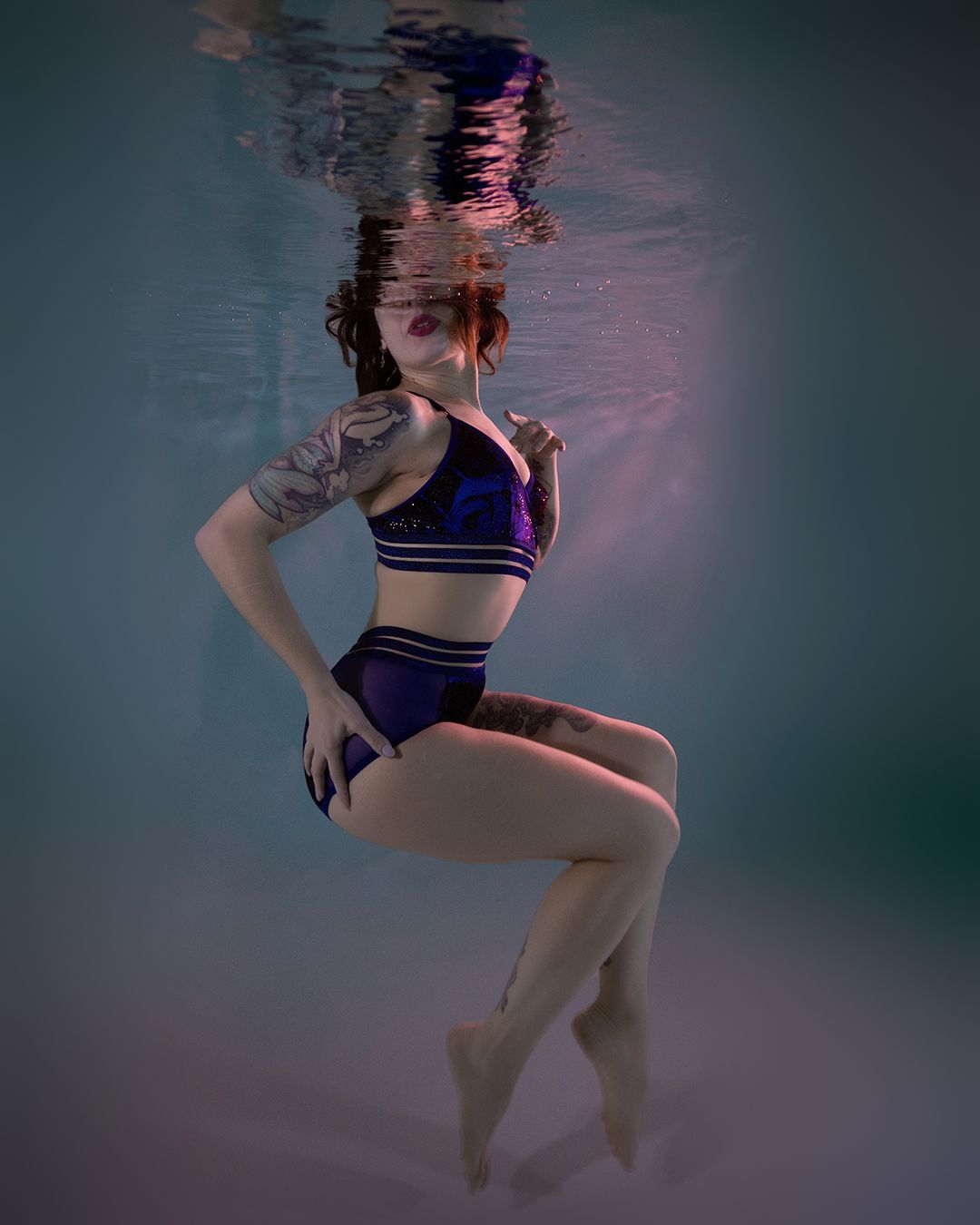 Woman in purple bikini posing gracefully underwater in a pool with teal-toned water and light reflections.