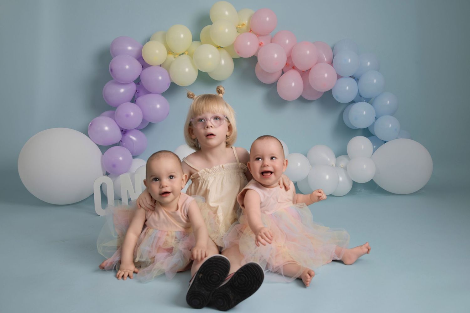 Little children sit beneath a pastel balloon arch in matching soft pink outfits against a light blue backdrop.