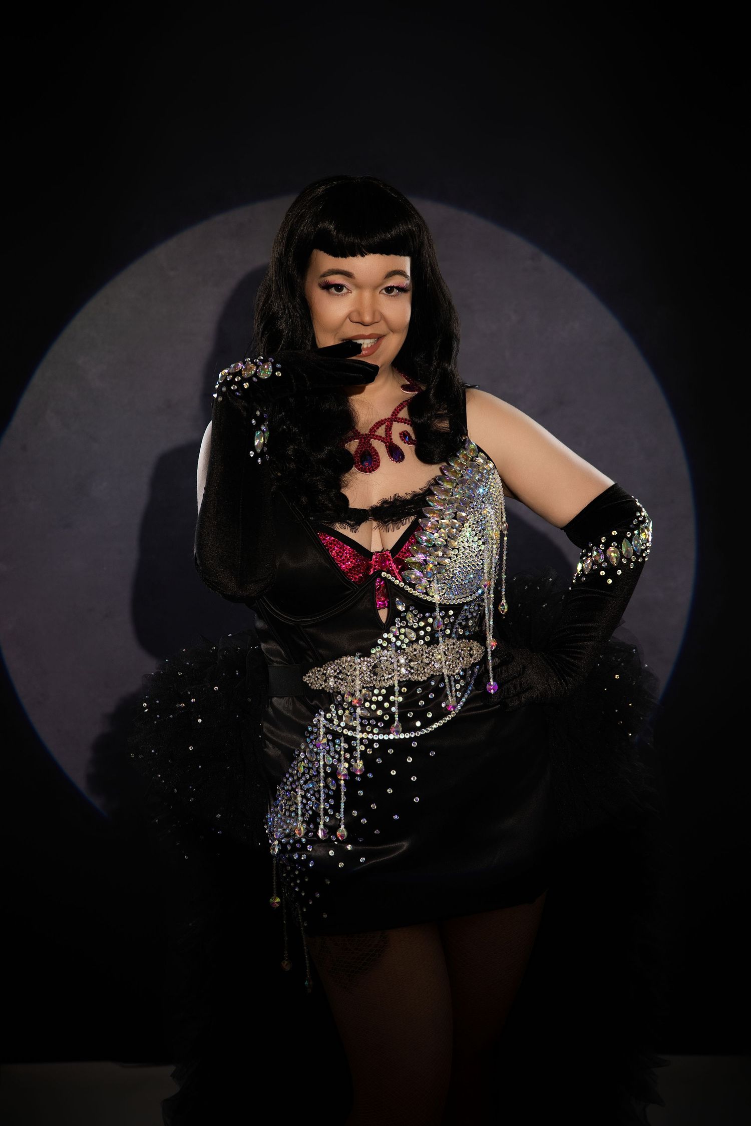A performer in a sparkling black burlesque costume with rhinestones poses dramatically against a dark spotlight background.