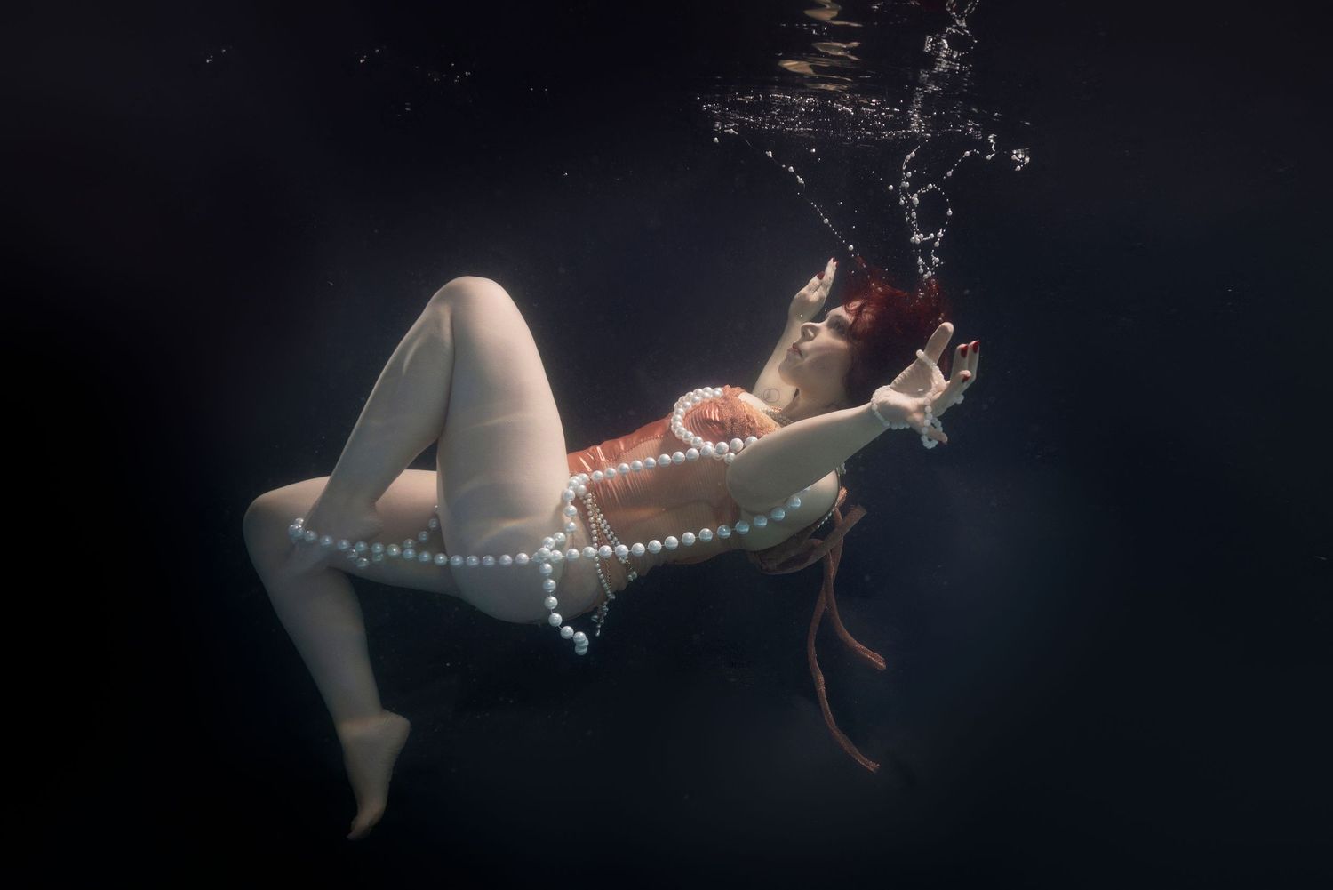 Red-haired woman underwater wearing pearl jewelry, gracefully floating against dark background.