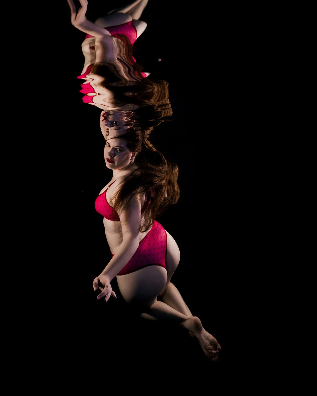 Woman in pink bikini poses gracefully underwater against a dark background, captured in a stunning artistic photography shot.