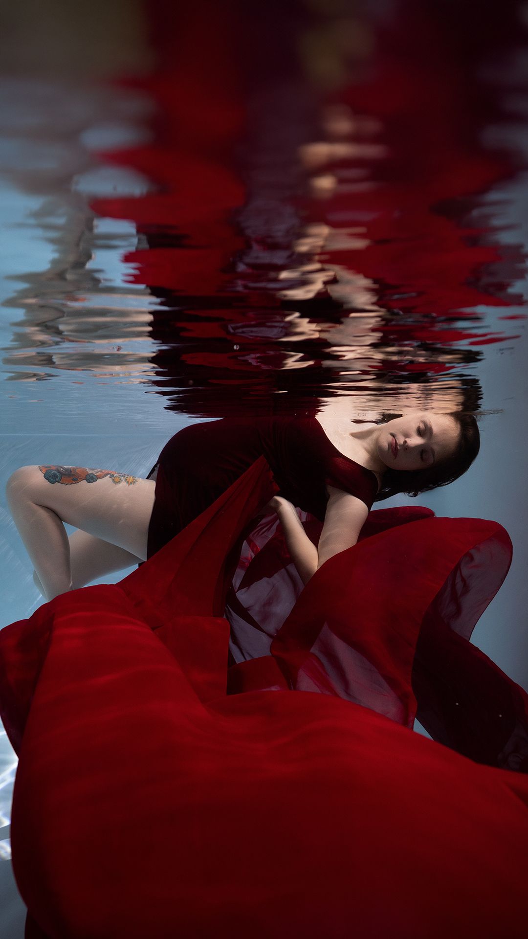 Woman in red dress floating underwater, red fabric billowing around her with reflection visible on water surface above.