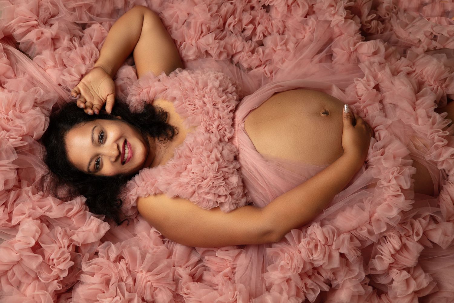 A person lying in a dreamy sea of pink tulle fabric wearing a soft pink dress for a maternity photo session.