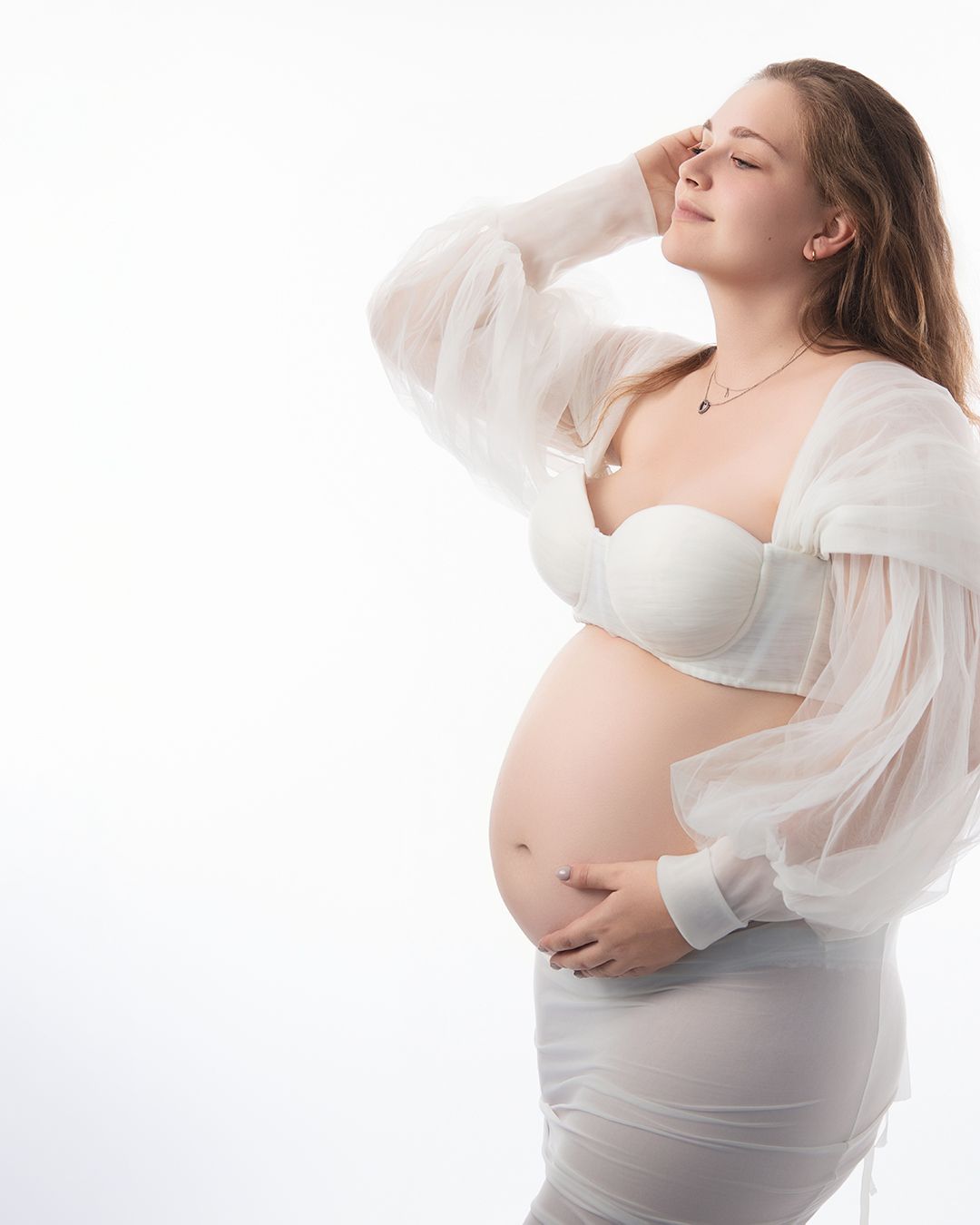 Pregnant woman in white sheer robe and bra cradling baby bump in elegant maternity studio portrait.